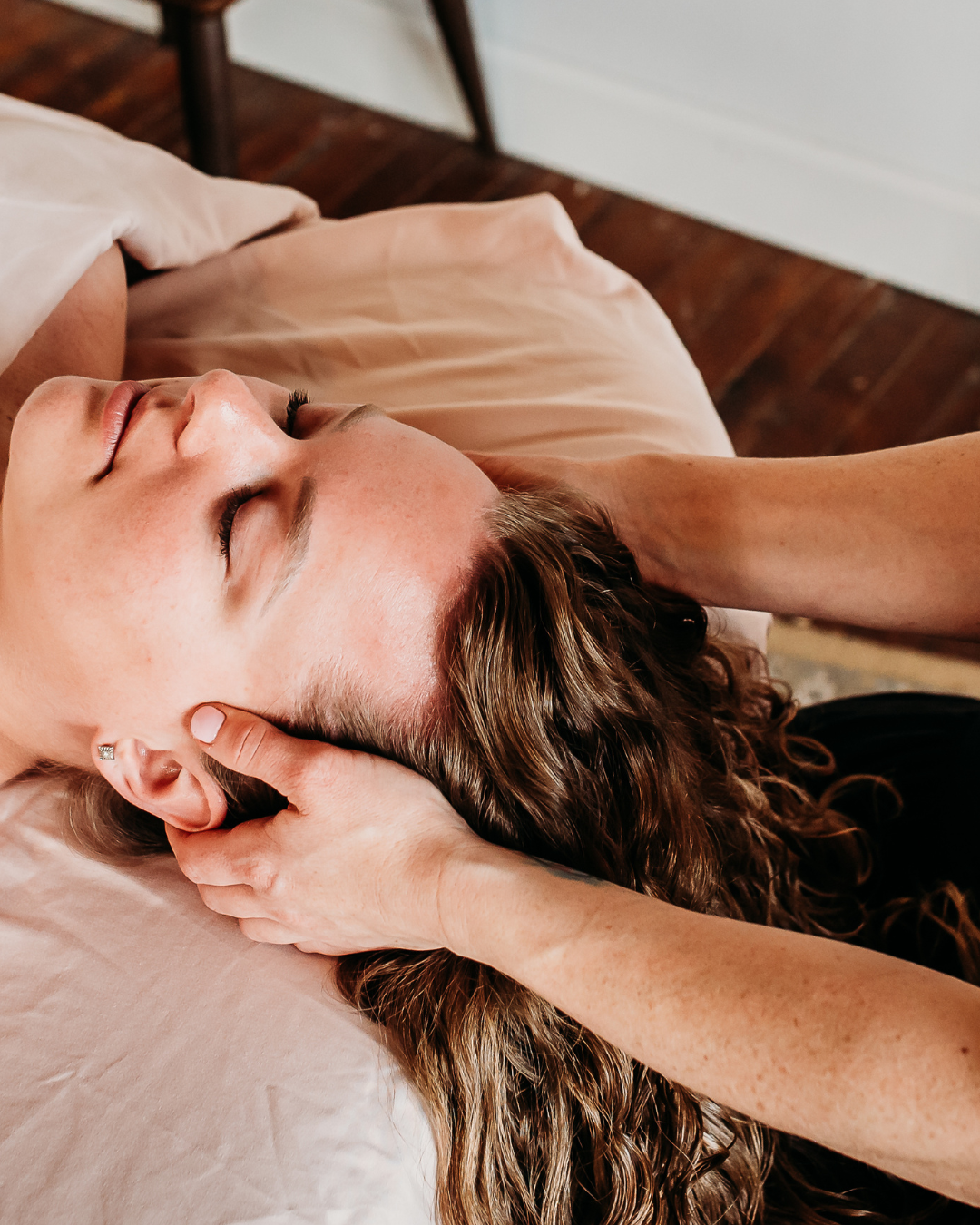 A woman receiving a neck massage on a massage table in a room with wooden flooring and furniture.
