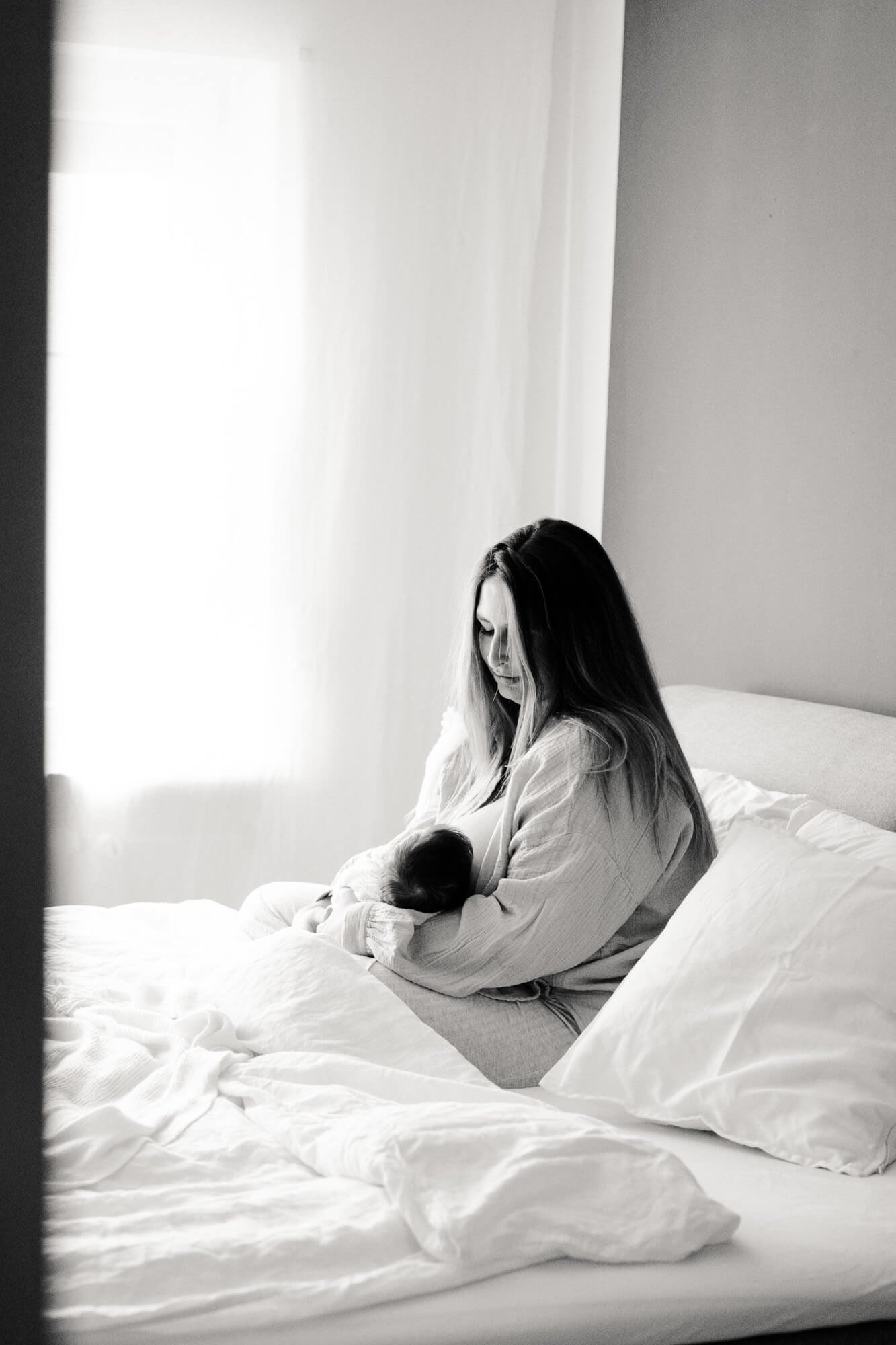 Madre sentada junto a la ventana con su bebé en brazos, ambiente íntimo y sereno.