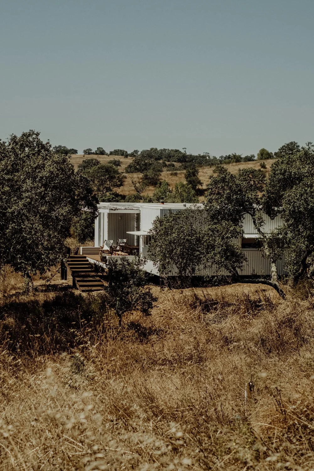 A white mobile home with a small deck and outdoor furniture, surrounded by trees and dry grassland, with rolling hills in the background under a clear sky.