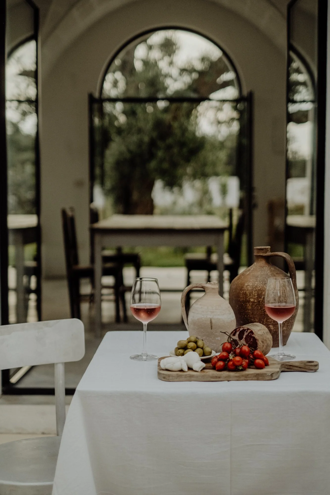 A table set with two glasses of rosé wine, two ceramic jugs, grapes, cherry tomatoes, and a wooden cutting board in a bright dining area with large windows and a view of a tree outside.