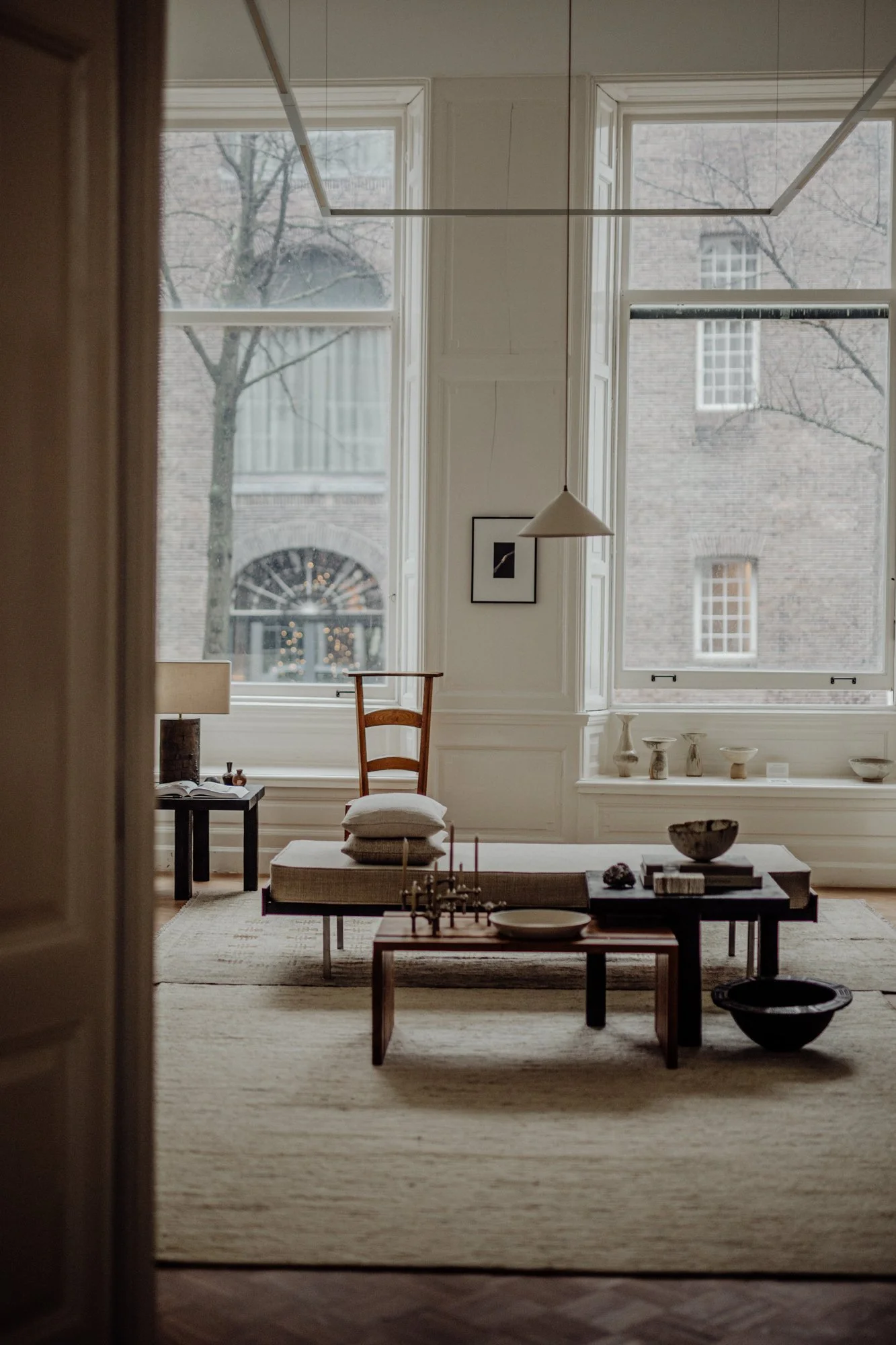 A minimalist living room with large windows, a wooden chair with pillows, a low coffee table with decorative items, and a pendant light hanging from the ceiling. The room features neutral tones and decorative pottery on a window ledge.