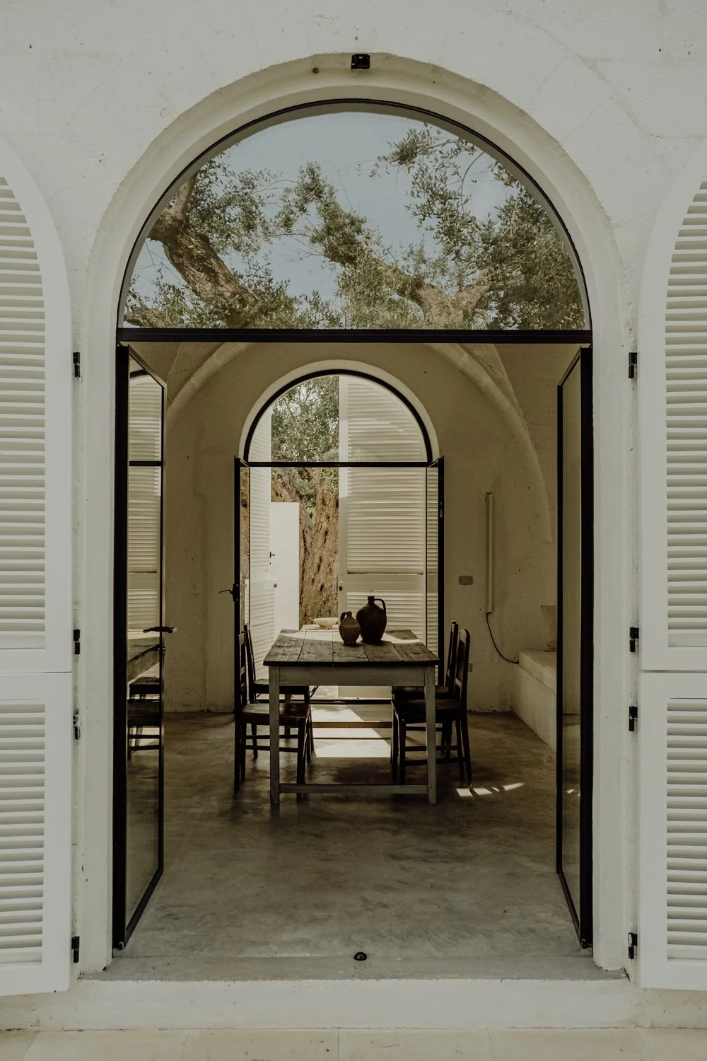 Open glass doors leading to a rustic dining area with a wooden table and chairs, overlooking a courtyard with trees and a clear sky.