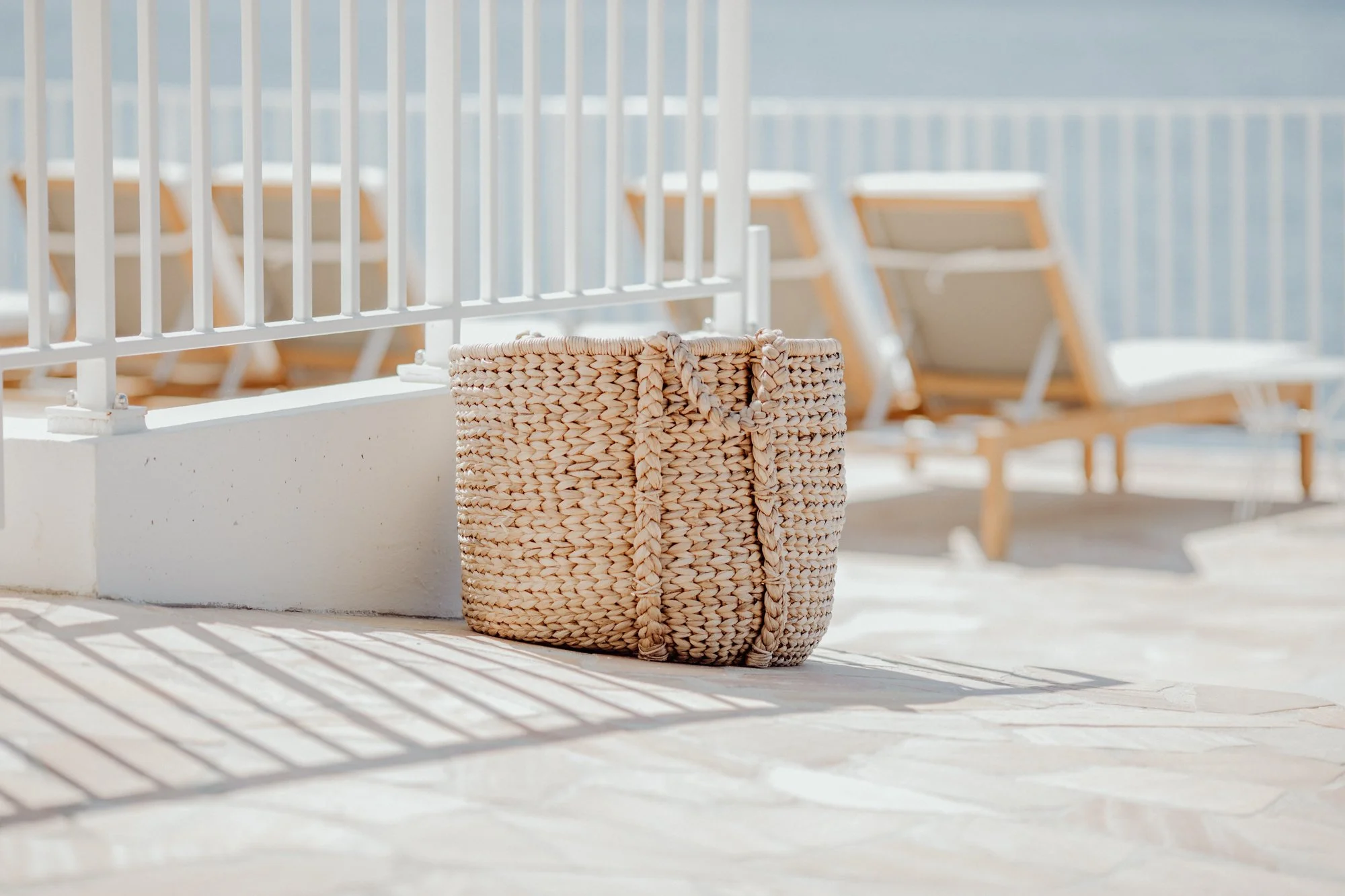 A woven straw bag resting on a tiled surface near a white railing, with lounge chairs in the background at a sunny outdoor poolside or beach area.