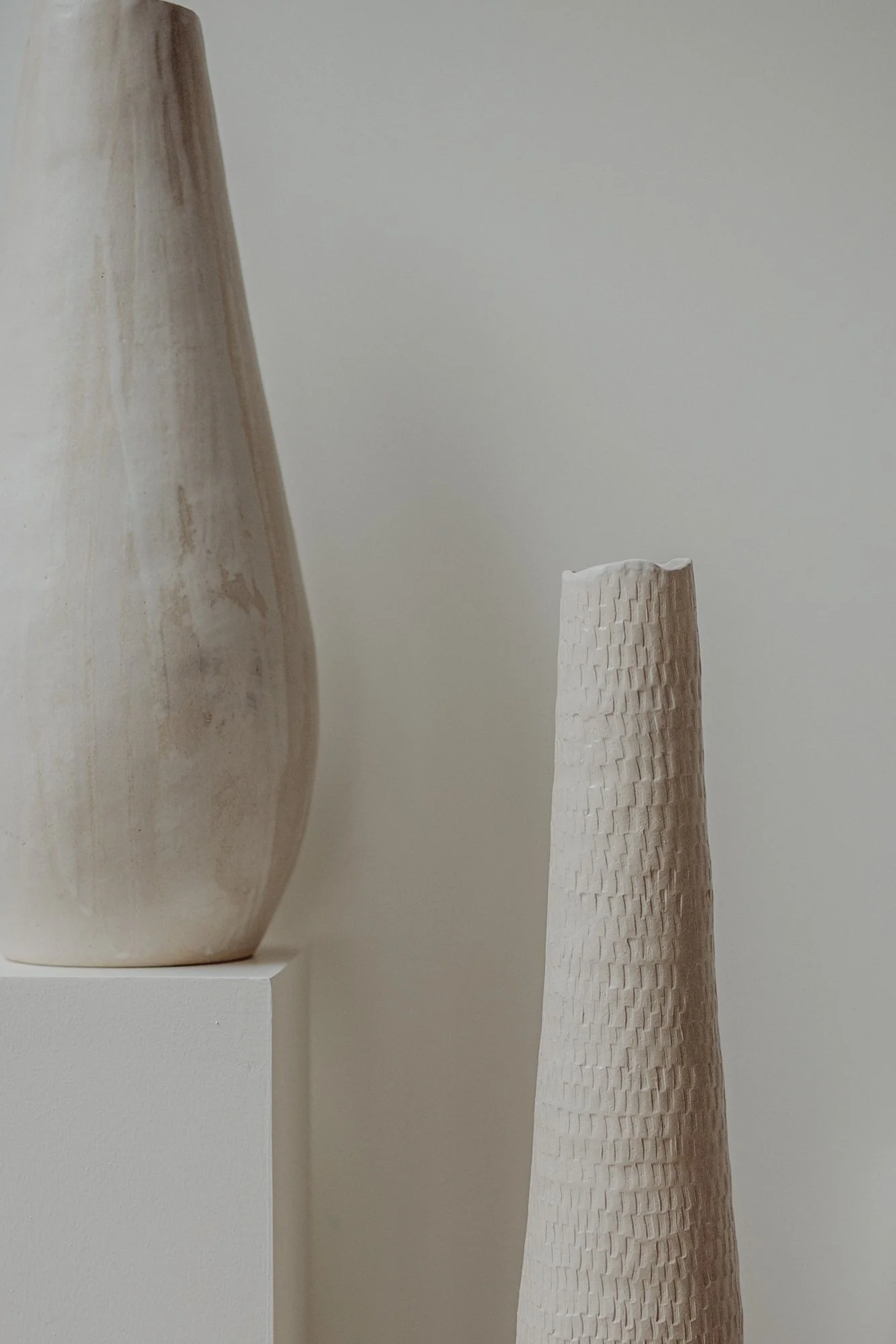 Close-up of two ceramic vases, one on a white pedestal and a textured one next to it, against a plain neutral background.