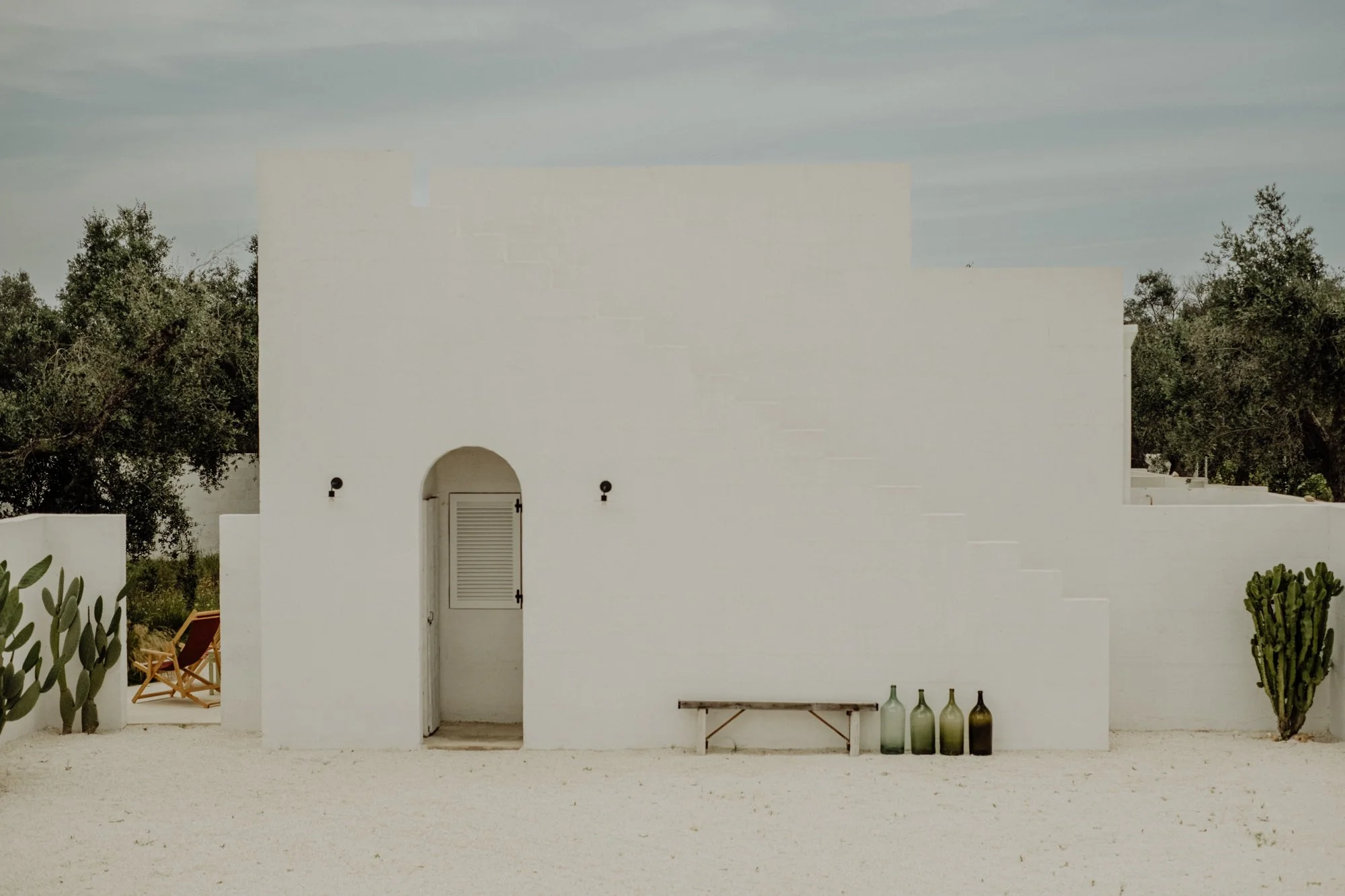 White building with small arched door and four glass bottles of varying shades of green and brown on the ground beside it, set against a natural background with trees and a cloudy sky.