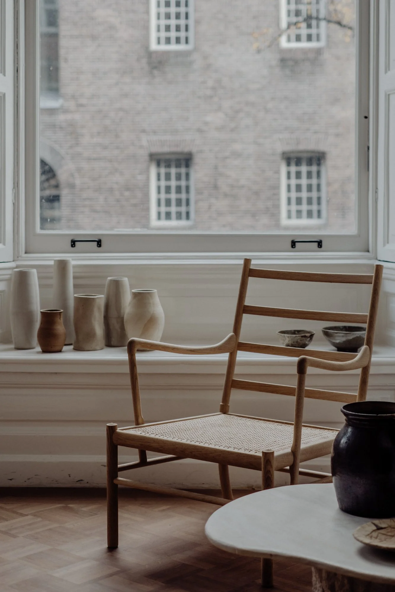Interior view of a room with a wooden chair with woven seat, a white table, and several ceramic vases on a windowsill. Outside the window, there is a brick building with multiple windows.