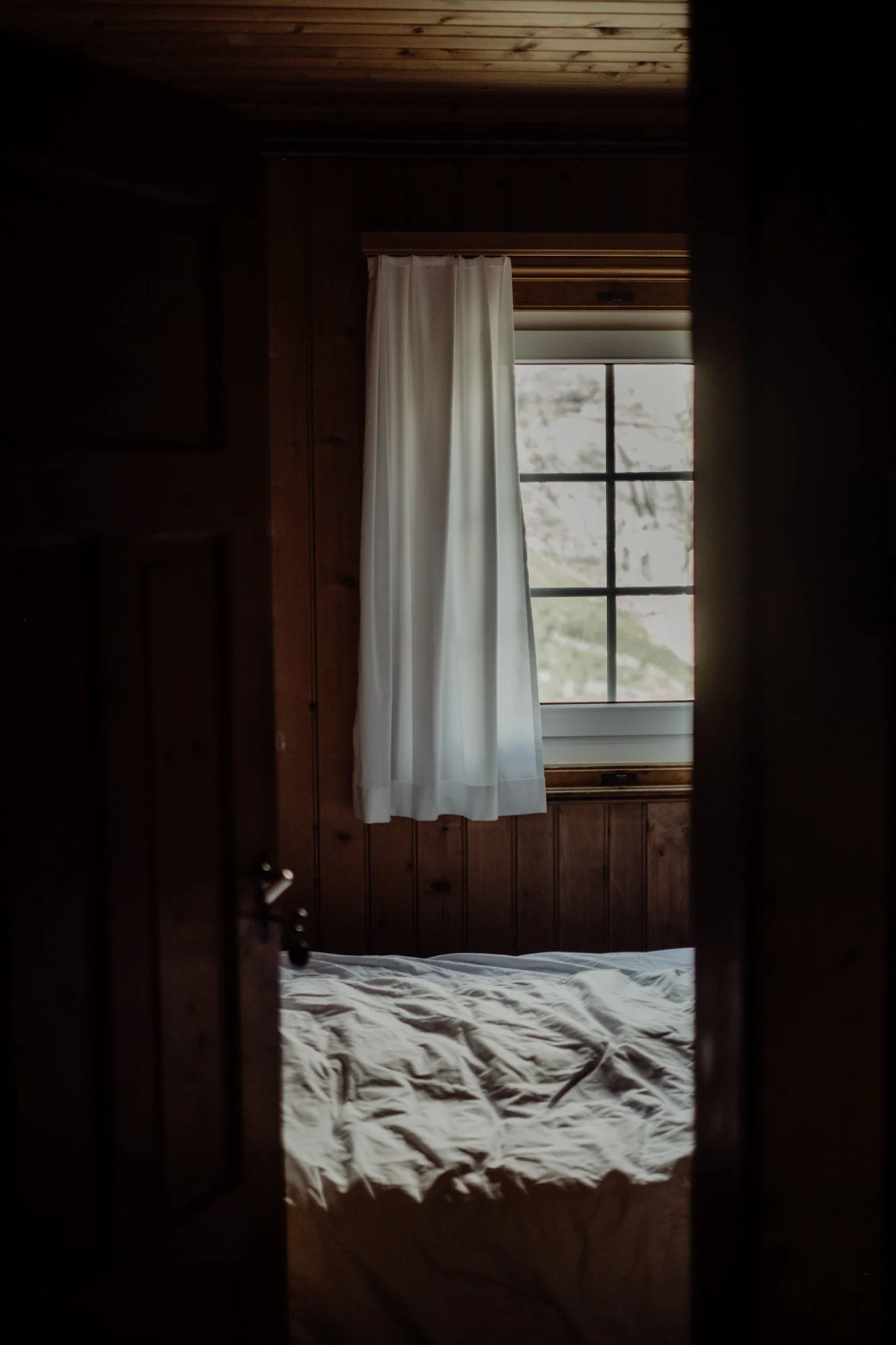View of a simple, rustic bedroom with a bed and a window with white curtains, wooden walls, and ceiling.