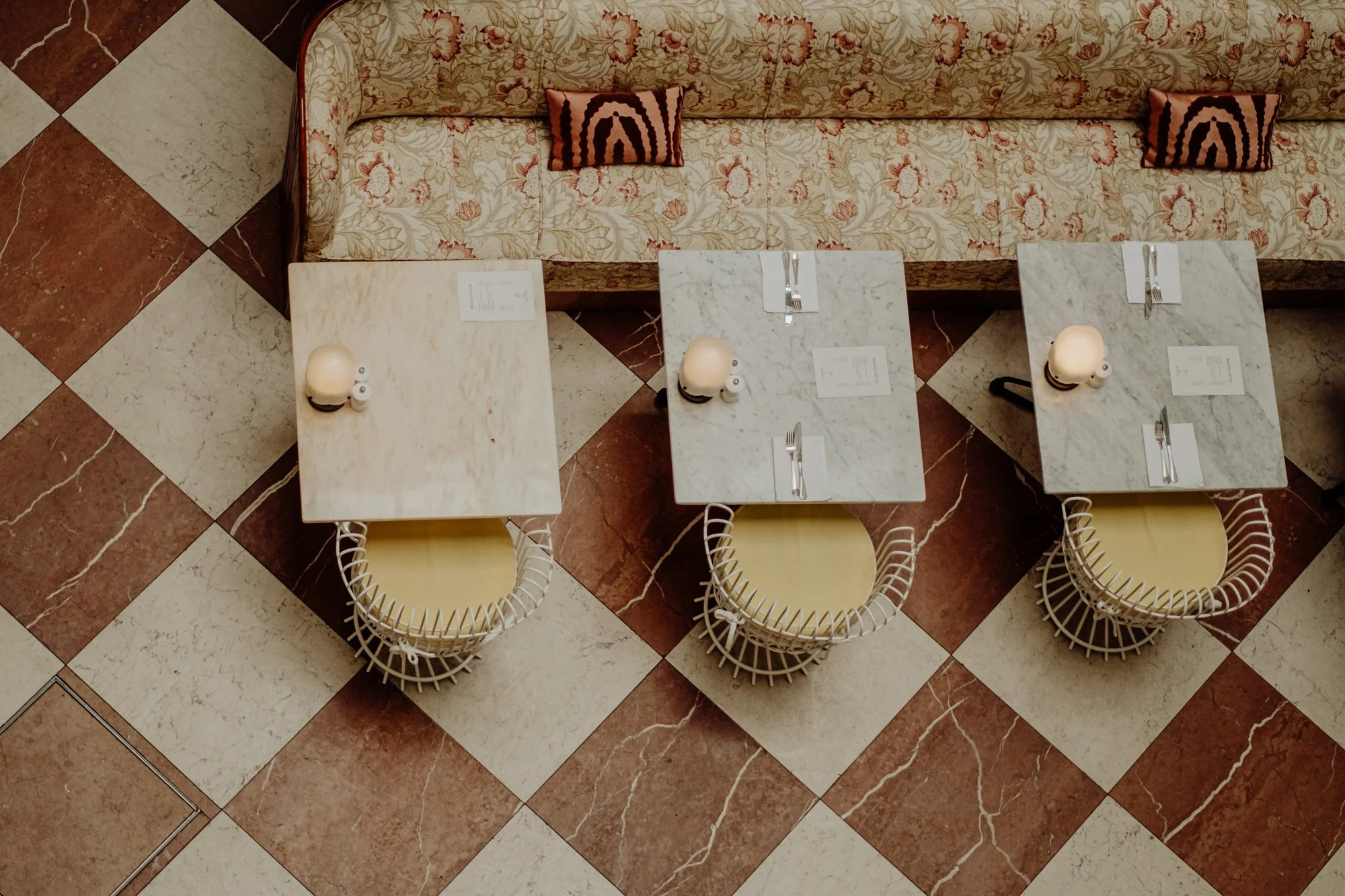 From above, three marble-topped tables are aligned in a row in front of a vintage floral-patterned sofa with pillows, on a checkered marble floor. Each table has a small lamp, salt and pepper shakers, and utensils.
