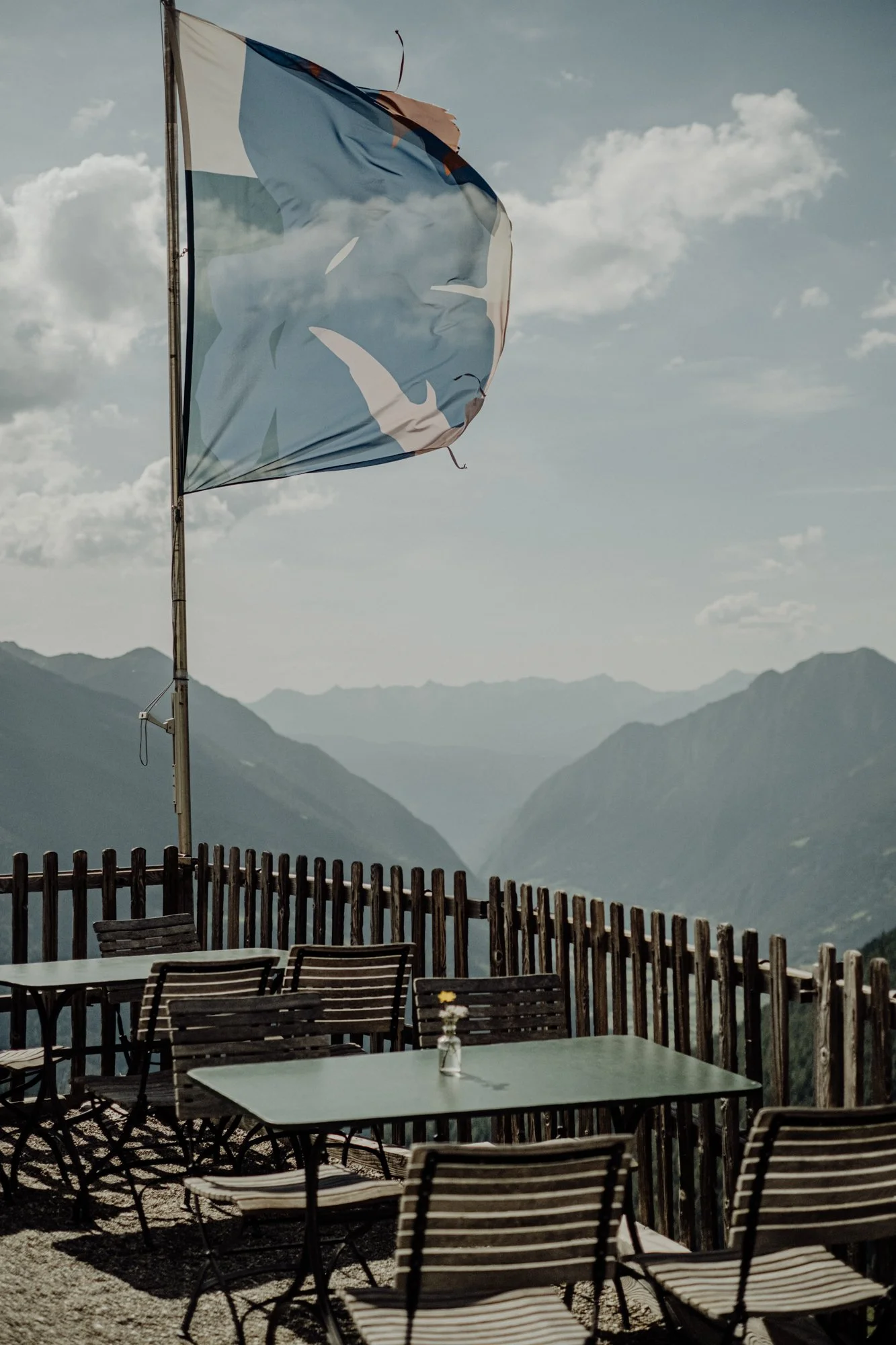 Outdoor patio with tables, chairs, and a mountain view in the background. A flag flaps in the wind against a partly cloudy sky.