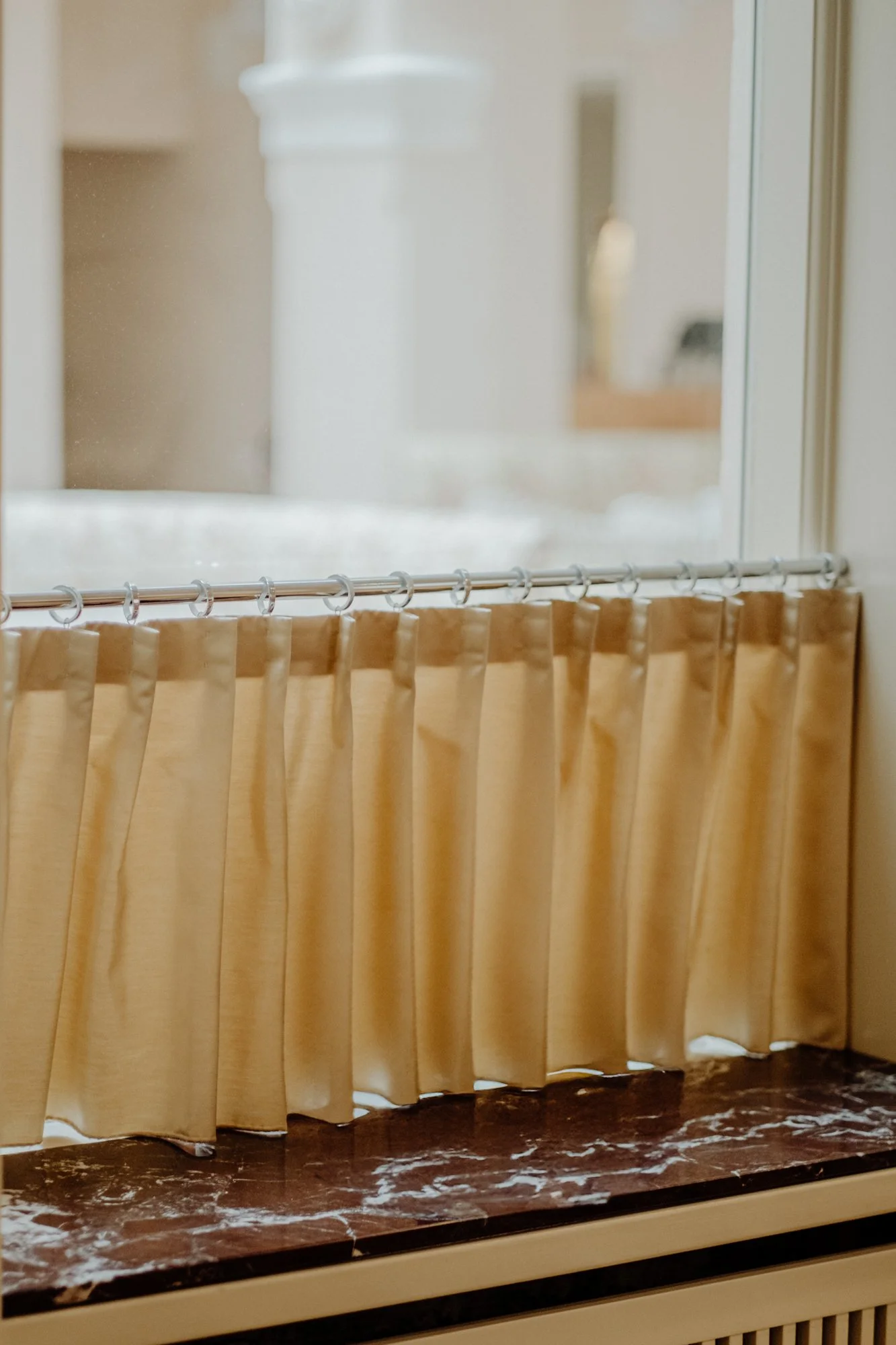 A bathroom window with closed beige curtains, a marble countertop with scattered white powder or flour, and a reflection of a bathtub in the window glass.