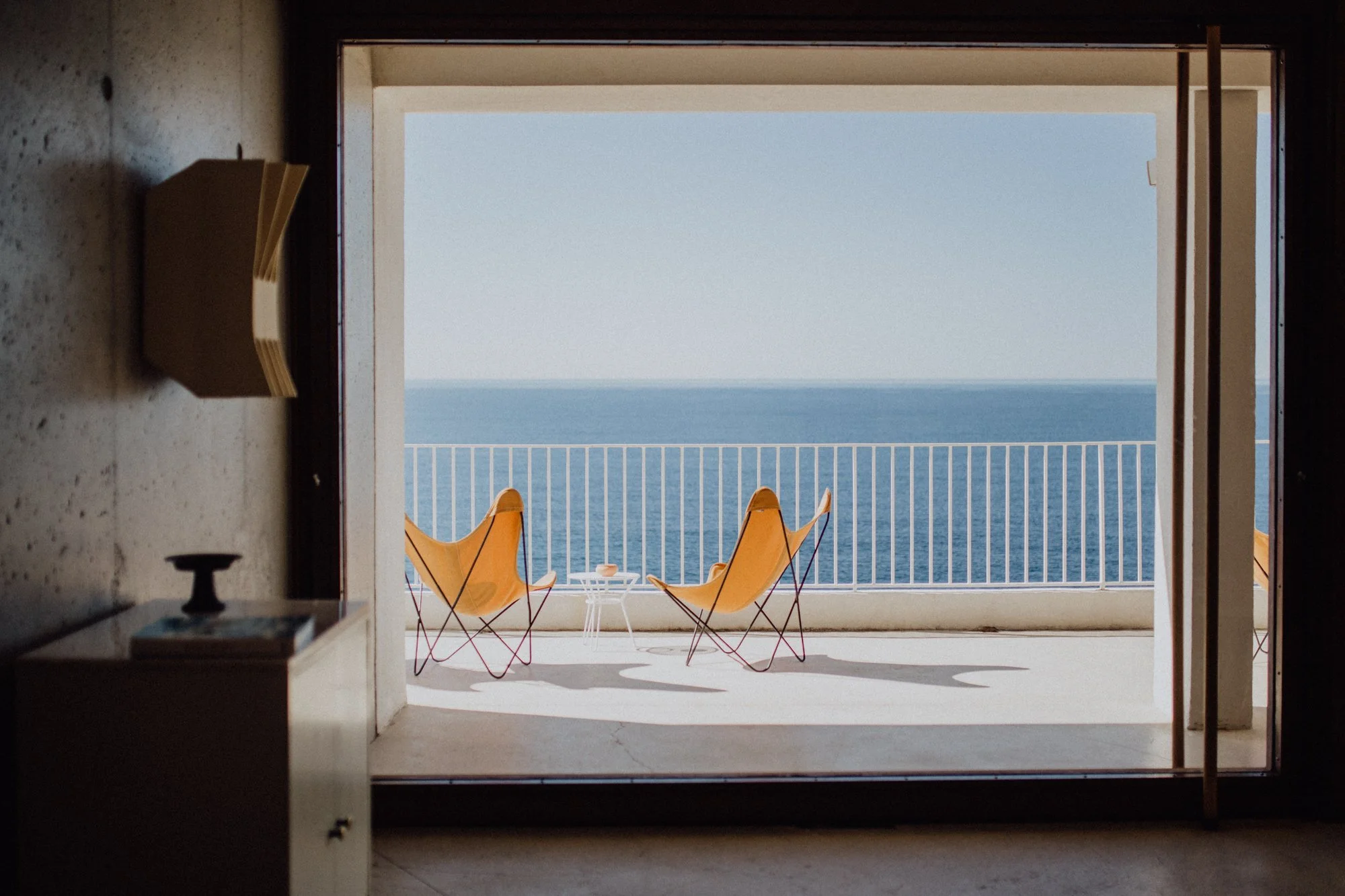 View from inside a room looking out onto a balcony with two yellow lounge chairs and a small white table, overlooking the ocean under a clear sky.