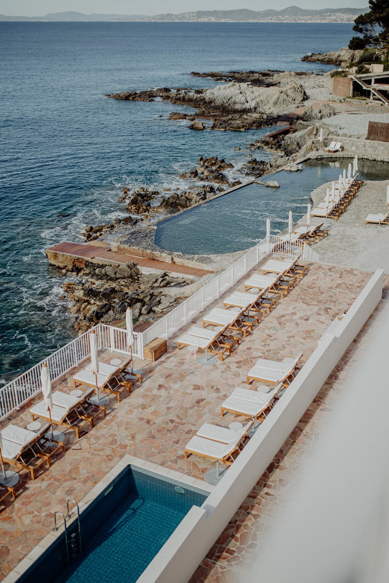A seaside pool area with lounge chairs and umbrellas overlooking the ocean, rocky shoreline, and distant hills.