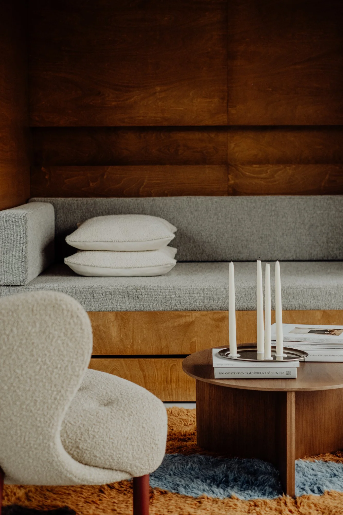 A cozy living room with a light beige armchair, a round wooden coffee table with white candles and magazines, a built-in gray sofa with white pillows, a wooden wall background, and a blue and orange rug.