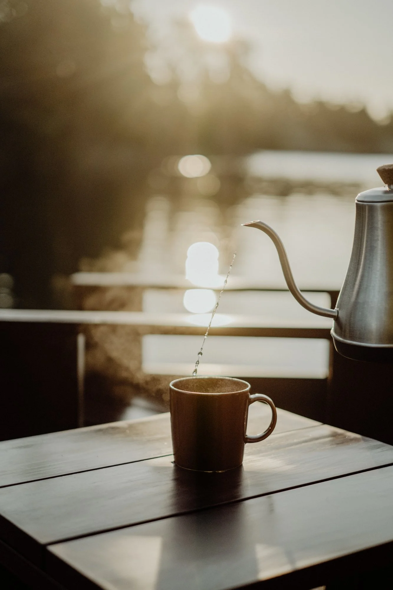 A warm-toned photo of a metal gooseneck kettle pouring hot water into a ceramic mug on a wooden table, with a blurred outdoor background and sunlight reflecting on the water.