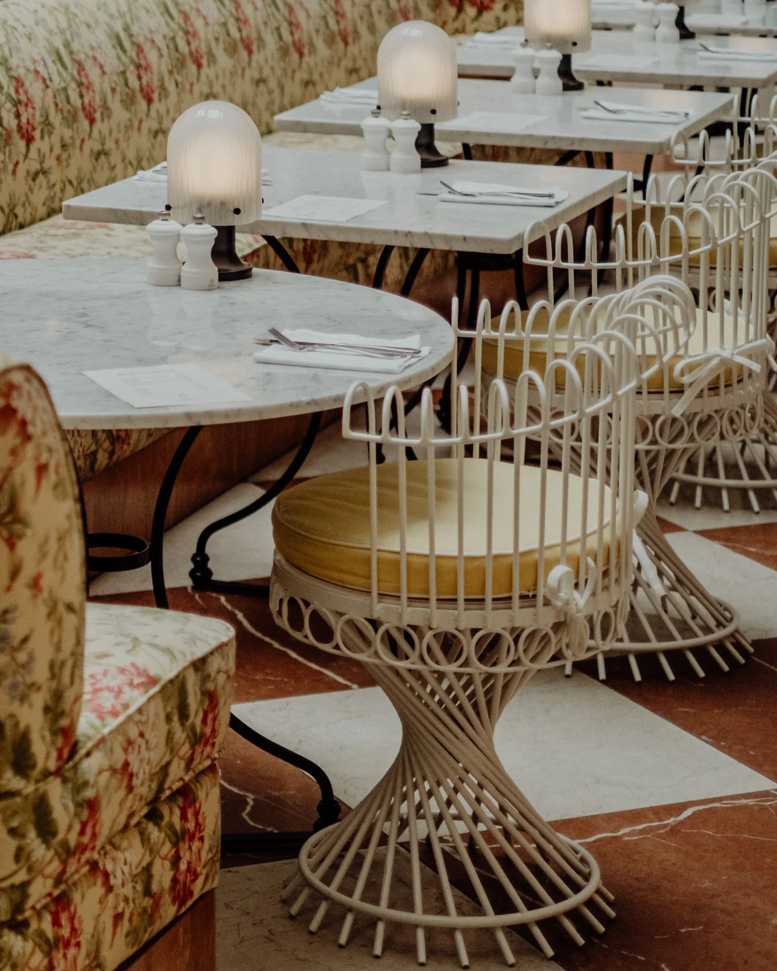 Empty restaurant tables with white marble tops, glass lamps, and white chairs with yellow cushions in a cozy, vintage-style dining area.