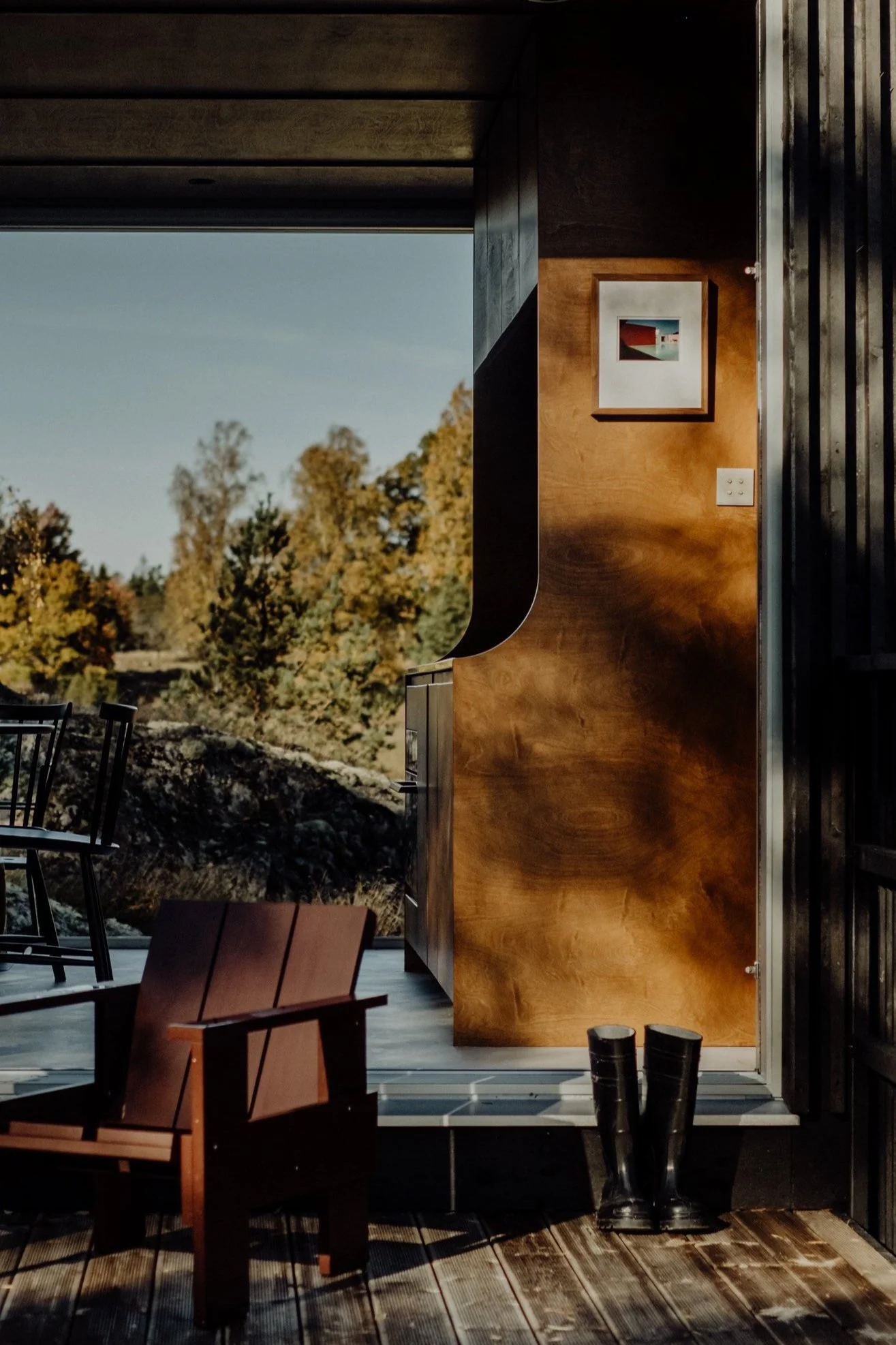 Interior view of a room with a wooden chair, pair of black rubber boots, and a wooden wall with a framed picture, overlooking a landscape with trees and rocks.