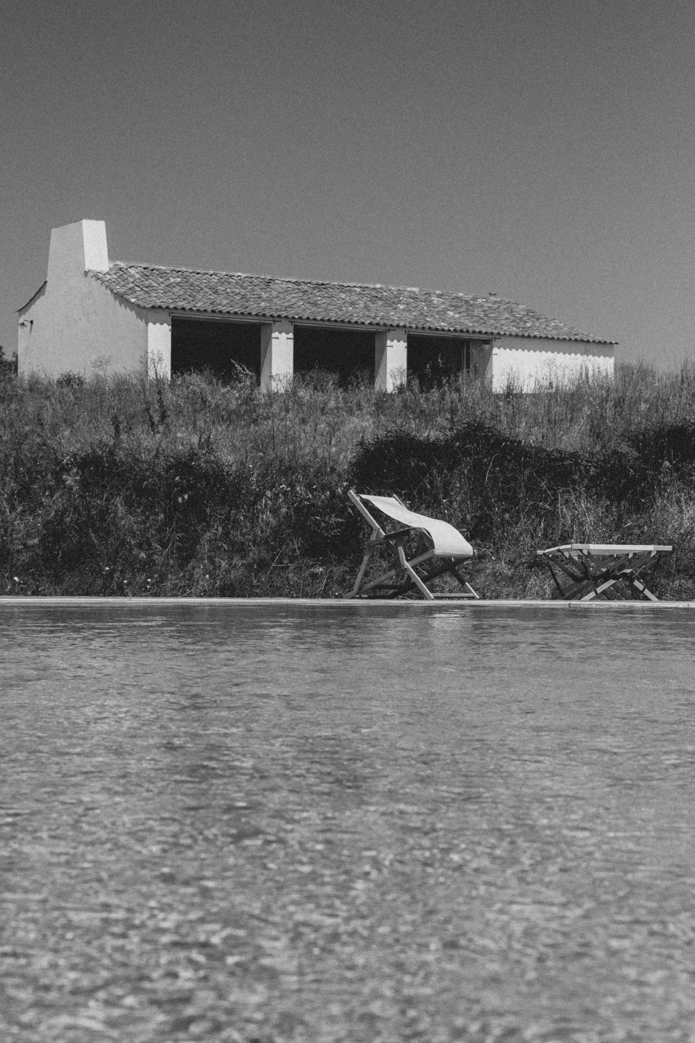 Black and white photo of an outdoor scene featuring a house with a tiled roof, a grassy hillside with shrubbery, and two lounge chairs near a body of water.