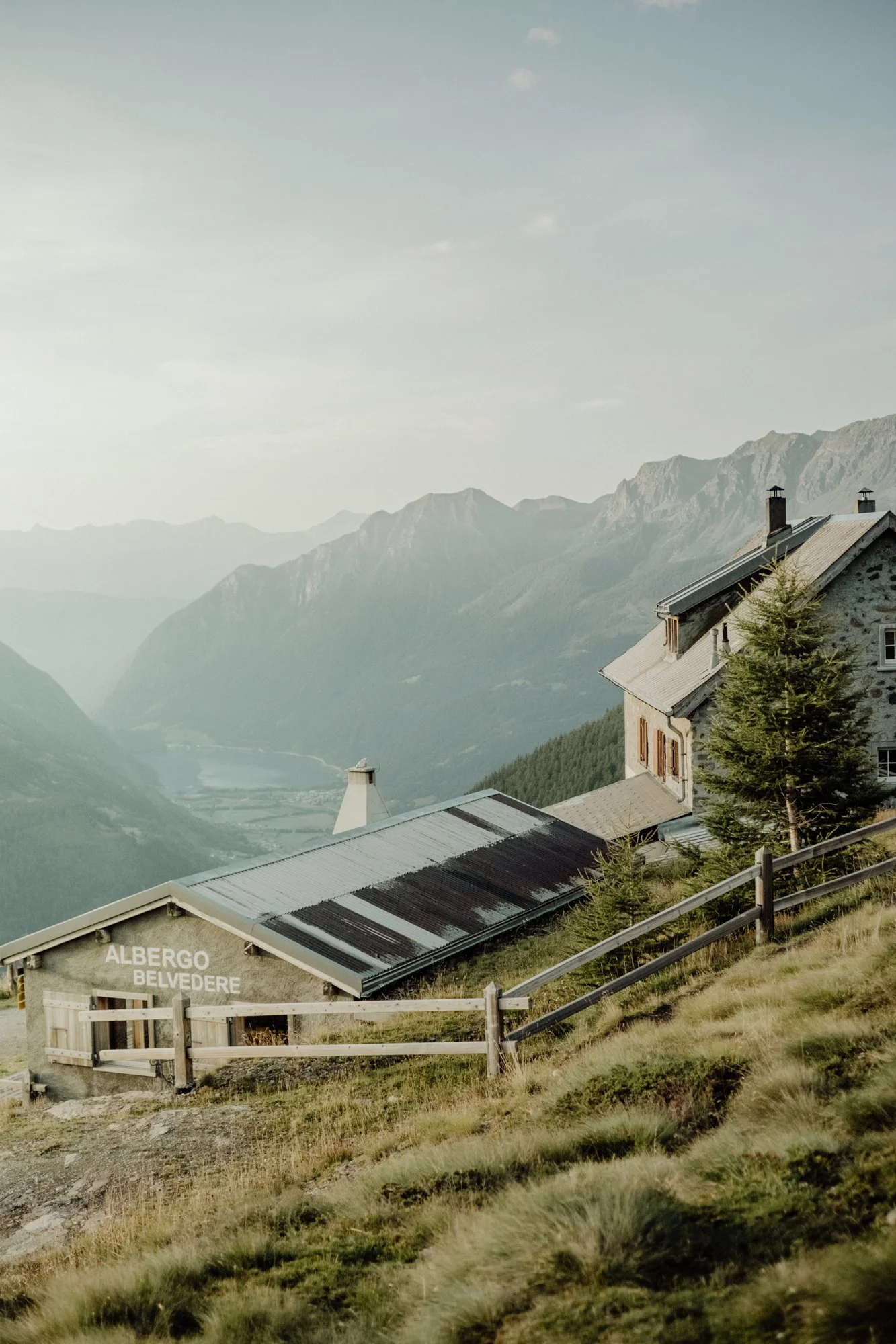 A scenic mountain landscape featuring a stone and wood building labeled 'Albergo Belvedere' with surrounding trees, sloped terrain, and distant mountains under a partly cloudy sky.