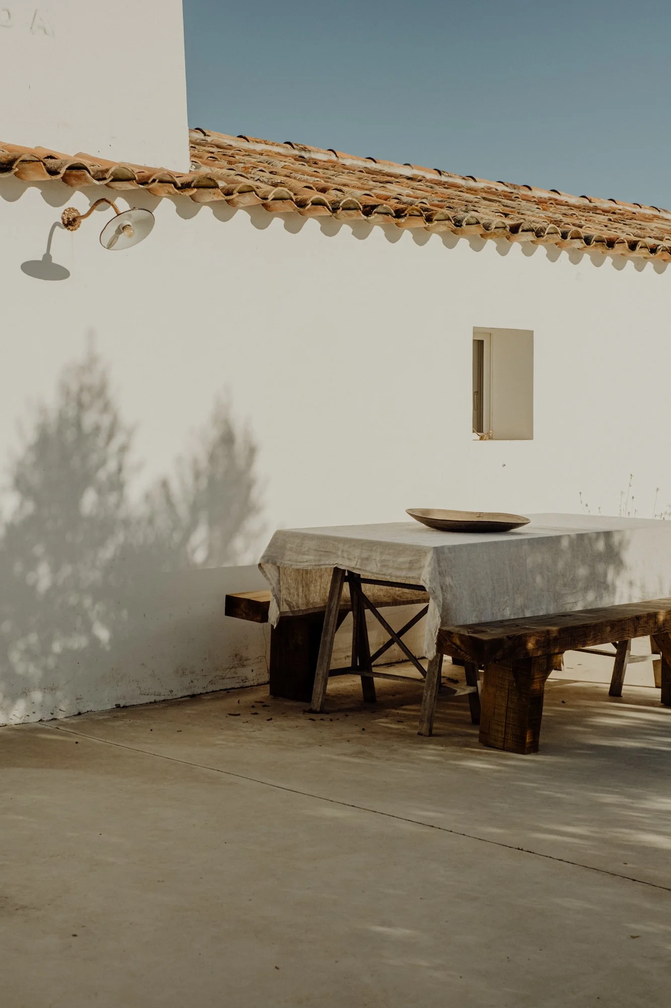 A white outdoor dining table with a beige tablecloth and a wooden bowl on top, surrounded by wooden benches, set against a white stucco wall with a small window. Shadows of trees are cast on the wall, and the scene is under bright daylight with a clear blue sky.