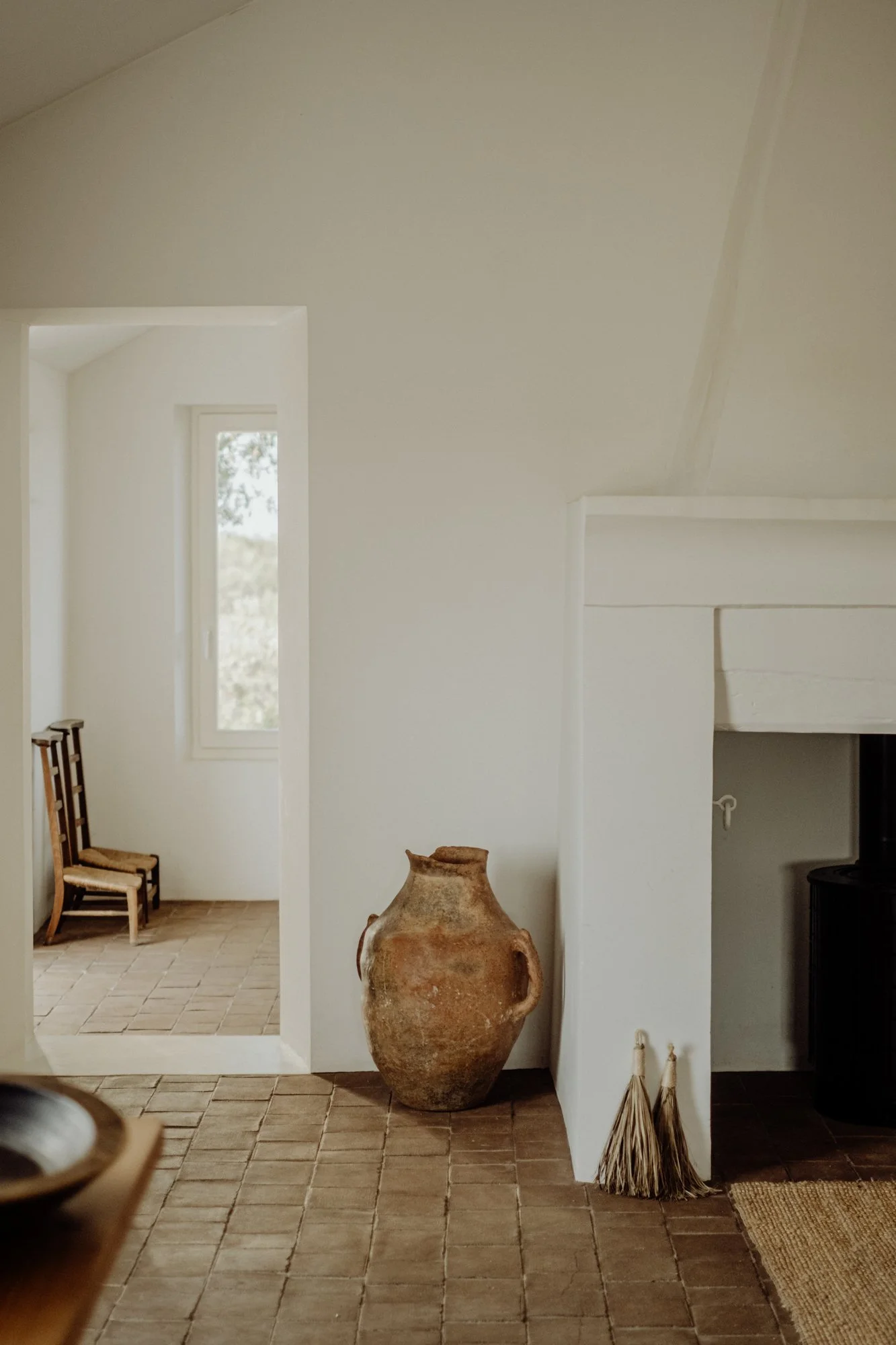 A rustic interior with a large ceramic vase, two small broom decorations, and a fireplace against white walls, with chairs and a window in the background.