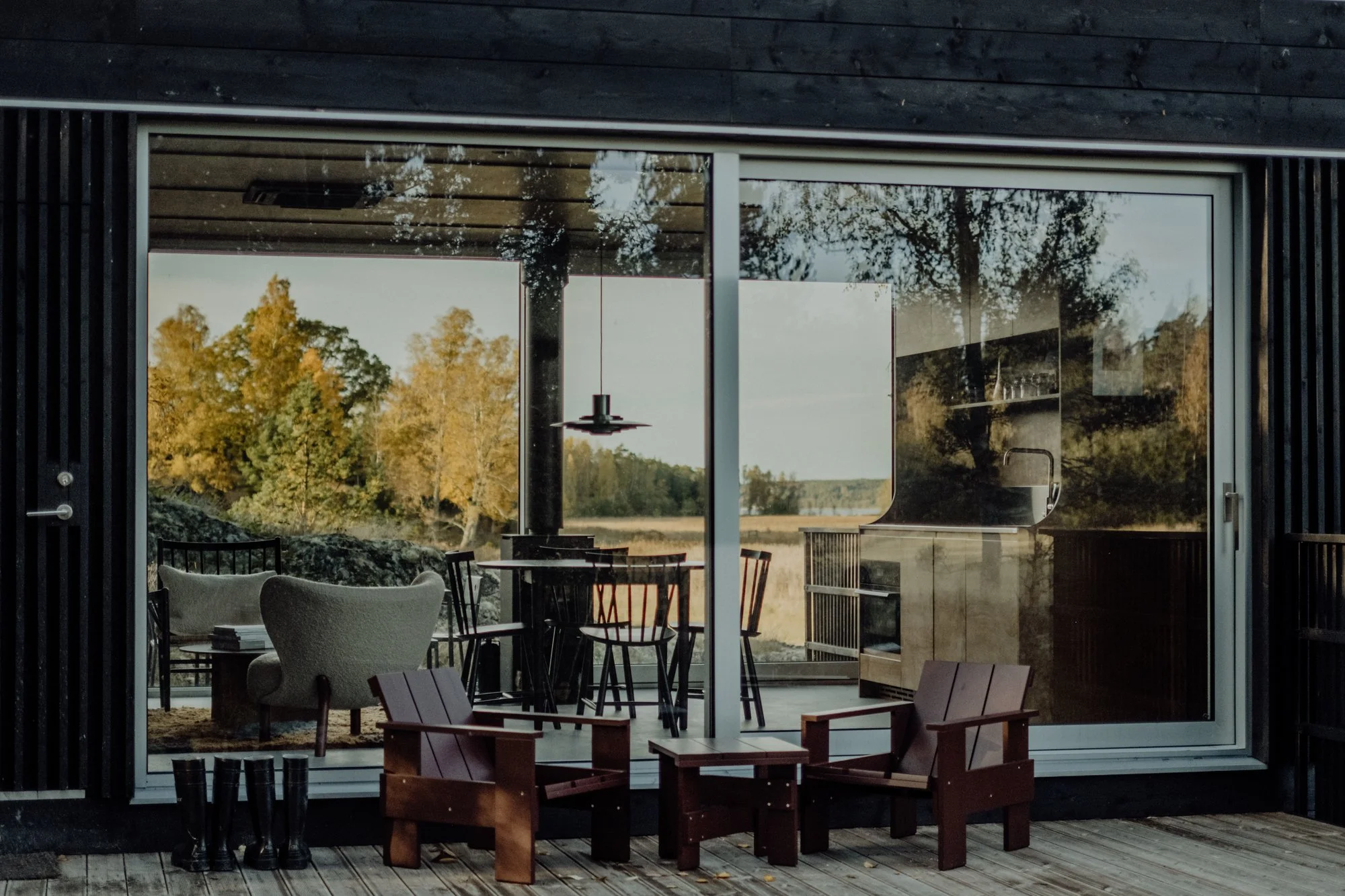 Outside view of a modern house with large glass sliding doors revealing a cozy interior with a dining table, chairs, an armchair, and a piano. The outdoor area has wooden decking, two small wooden chairs, and a picturesque view of trees with autumn foliage.
