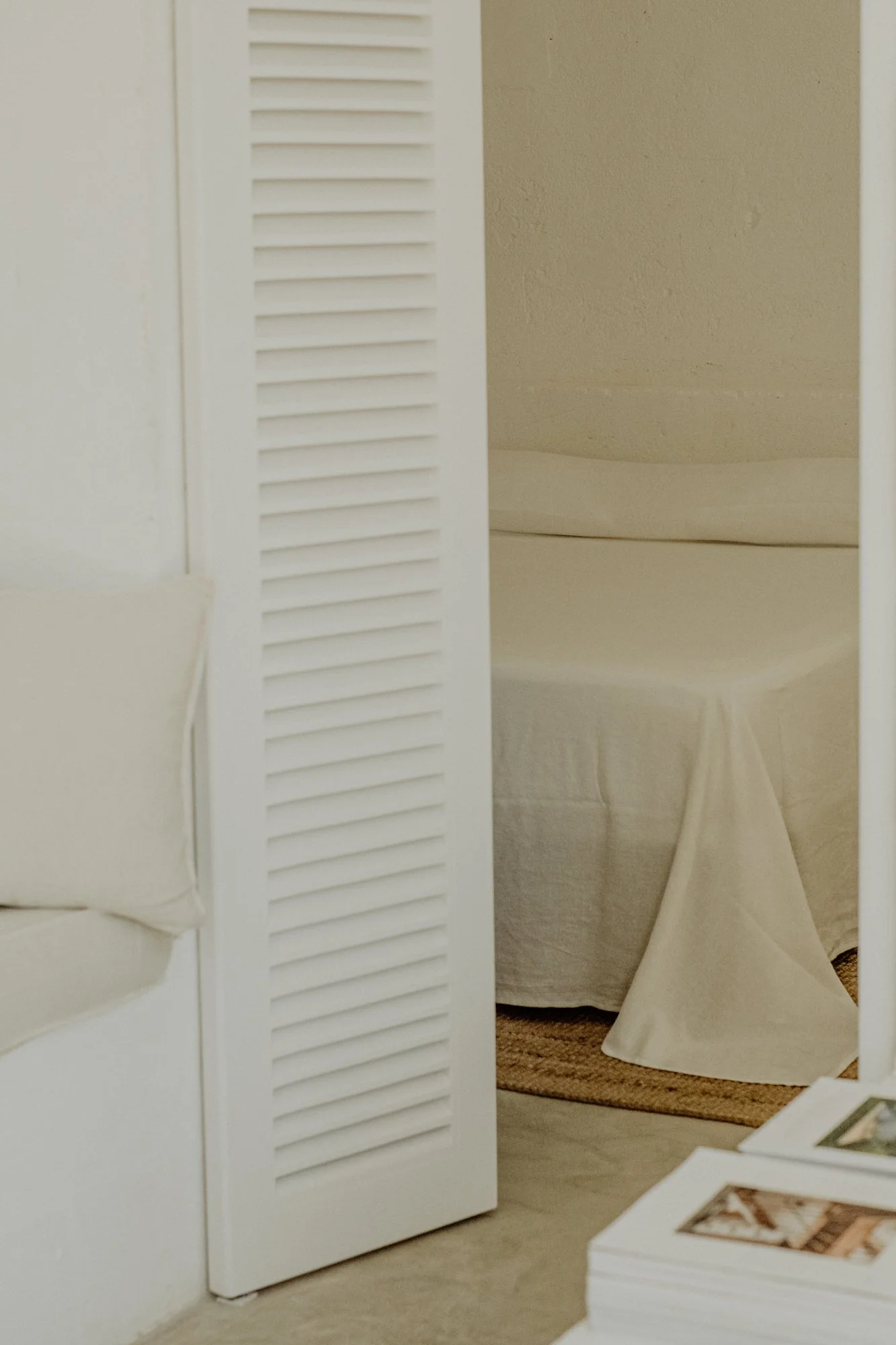 Partial view of a beige-colored room, showing a white louvered closet door, a section of a white sofa, a bed with a white bedcover, and a small table with photos.