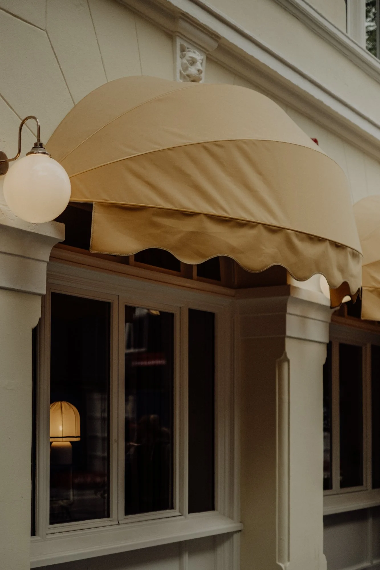 Close-up of a beige fabric awning above a window on a building exterior, with a white wall and an exterior light fixture nearby.