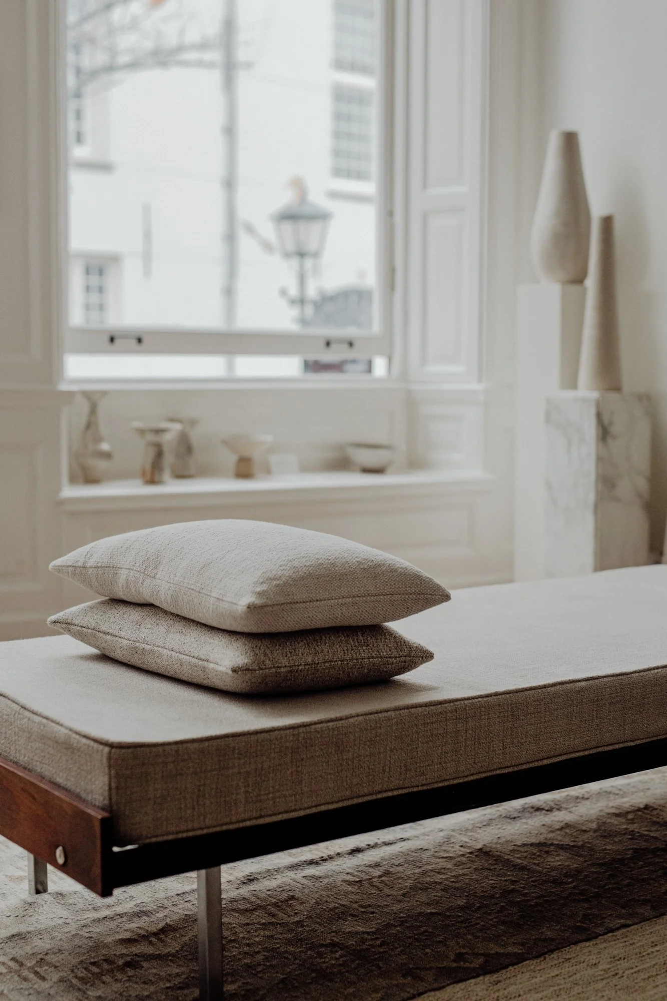 A minimalistic, neutral-colored room with a beige padded bench or daybed, two stacked pillows on top, a window with open shutters, and decorative vases and bowls on a ledge and shelves that add to the tranquil, modern aesthetic.