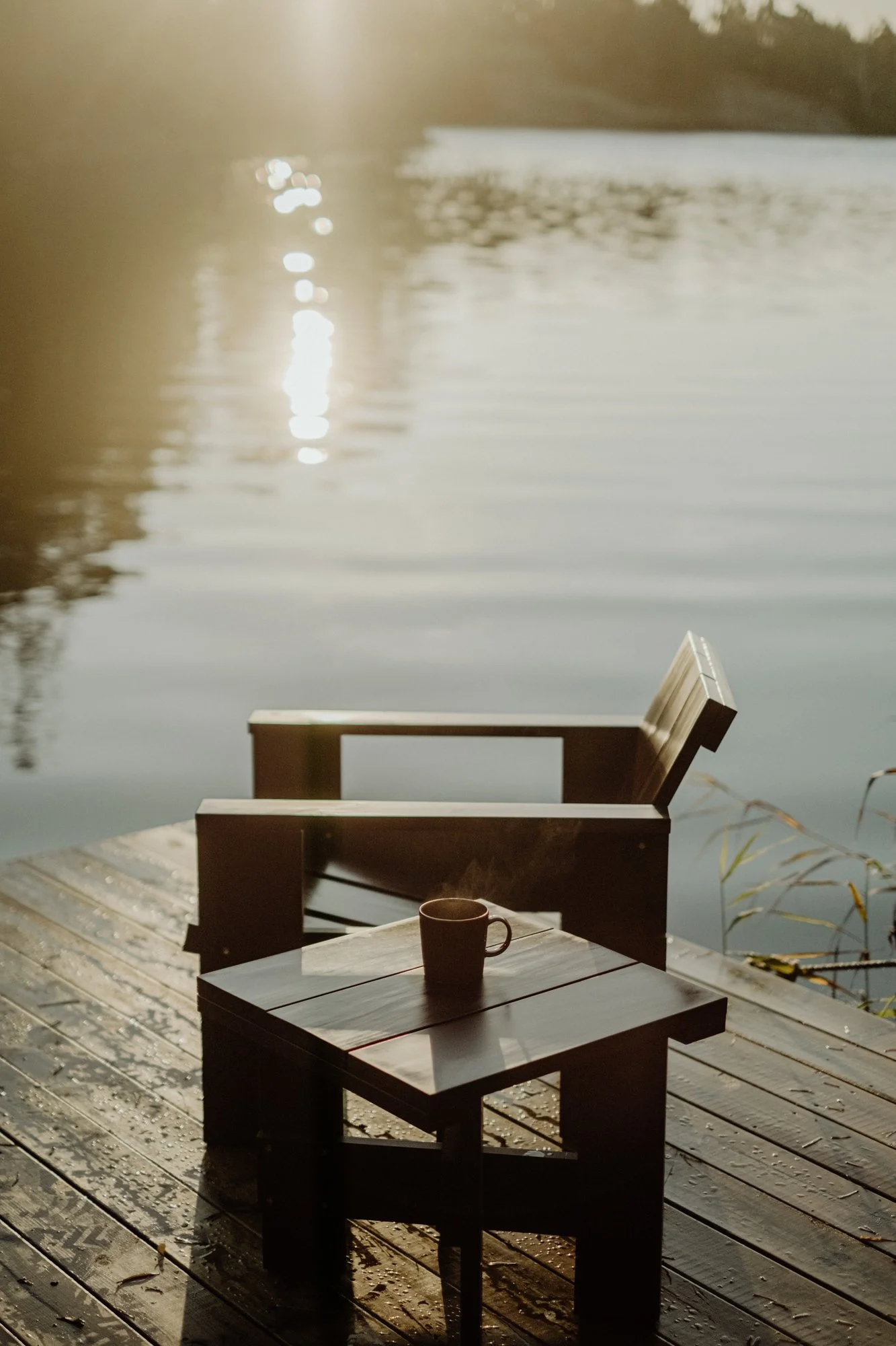 A small wooden table with a coffee mug on it, positioned on a wooden dock by a calm river with reflections of trees and sunlight on the water.
