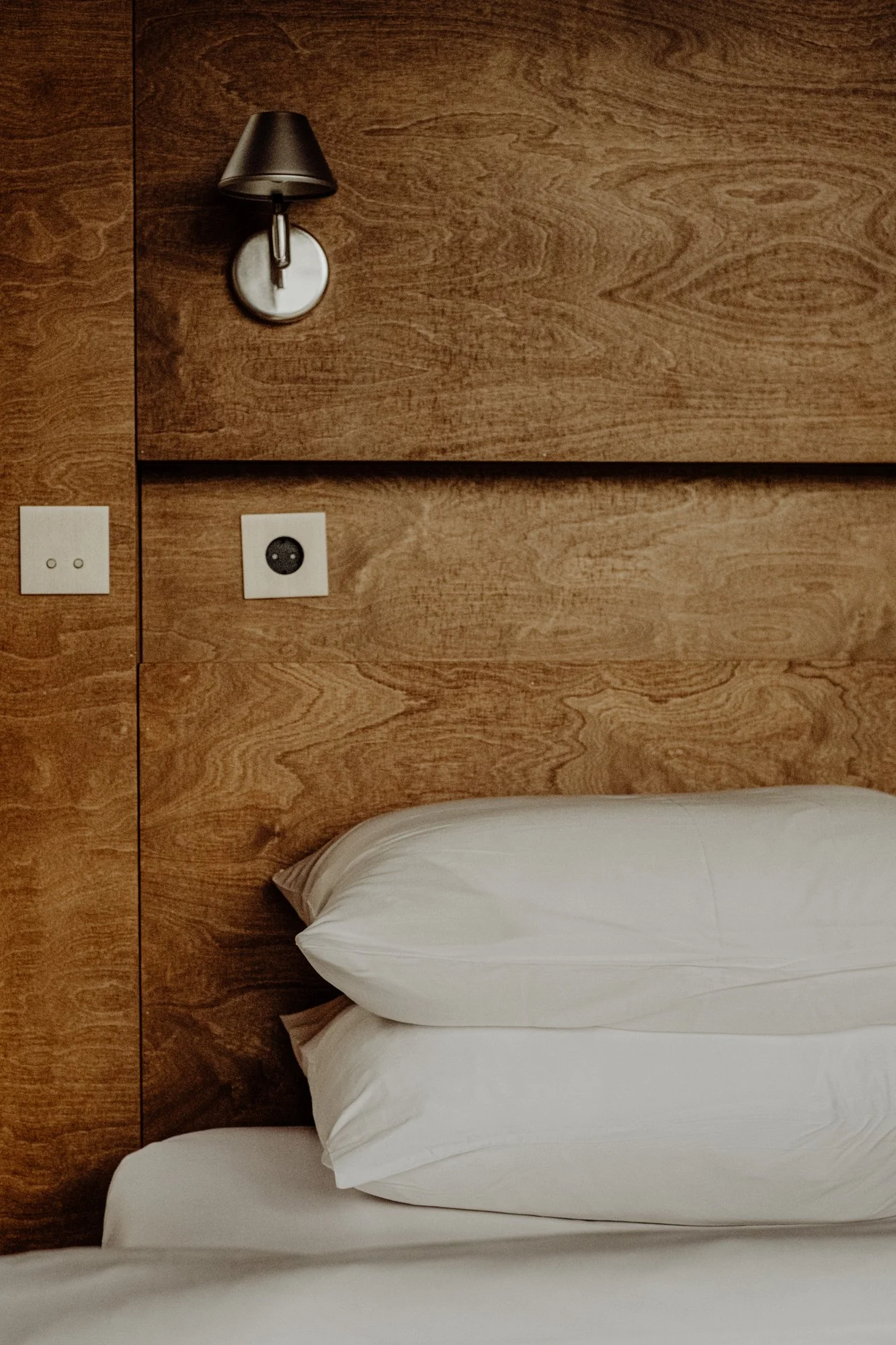 Close-up of a portion of a hotel bed with pillows, a wooden headboard, and a wall-mounted lamp. The wall has electrical outlets and a power socket.