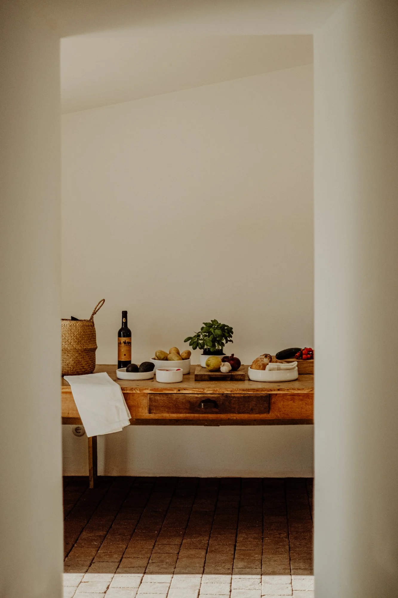 View of a rustic kitchen counter with various fruits, vegetables, a bottle of wine, and a potted plant, seen through a doorway.