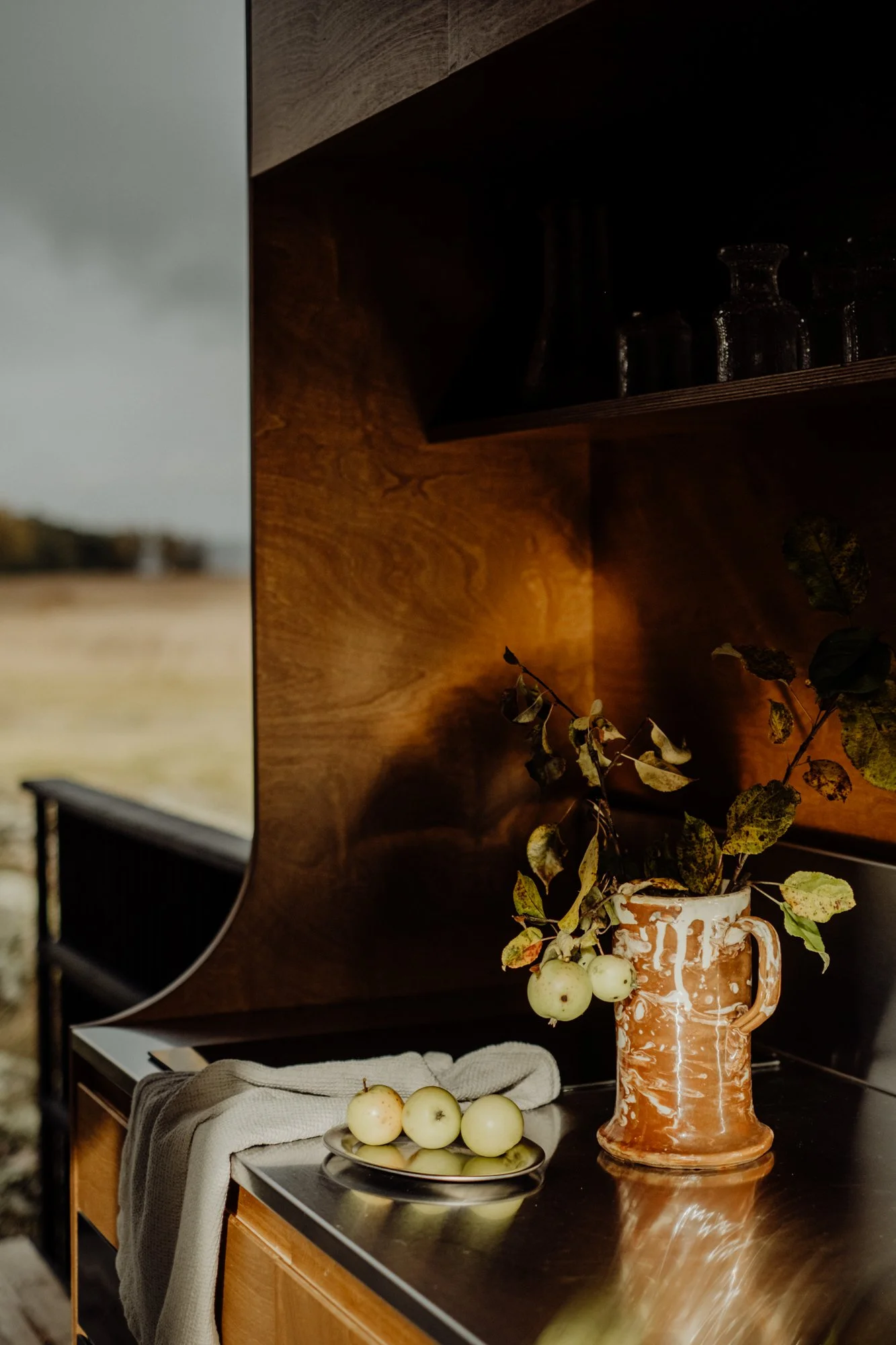 Still life image of a ceramic pitcher with leafy branches and apples on a kitchen countertop near a window with an outdoor field view.
