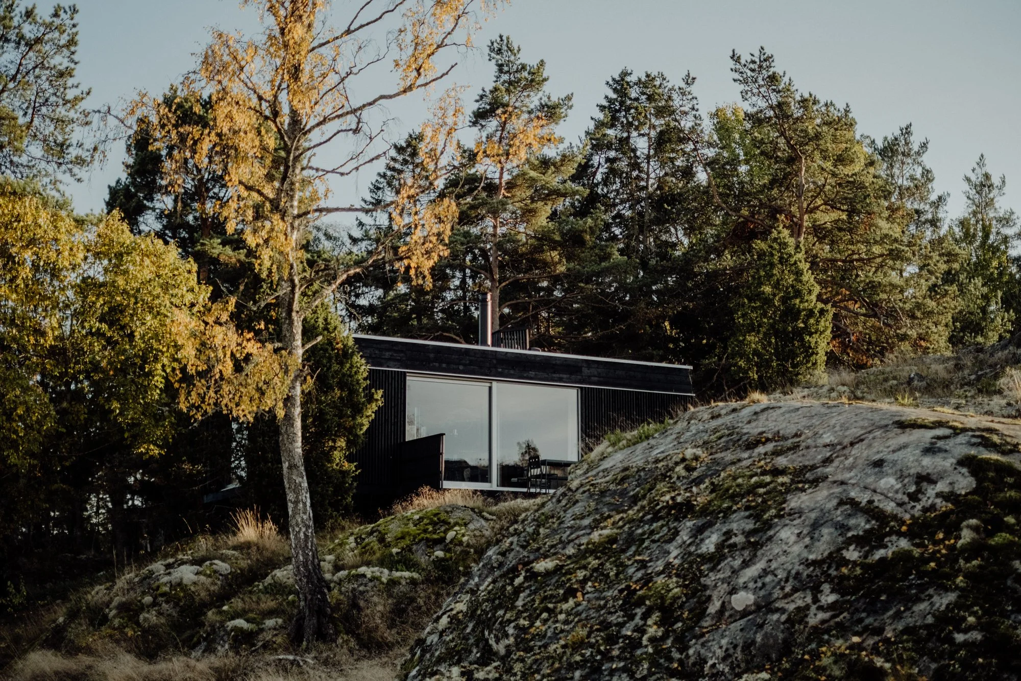 A modern house built on a hillside surrounded by trees and large rocks, with glass sliding doors and a patio.