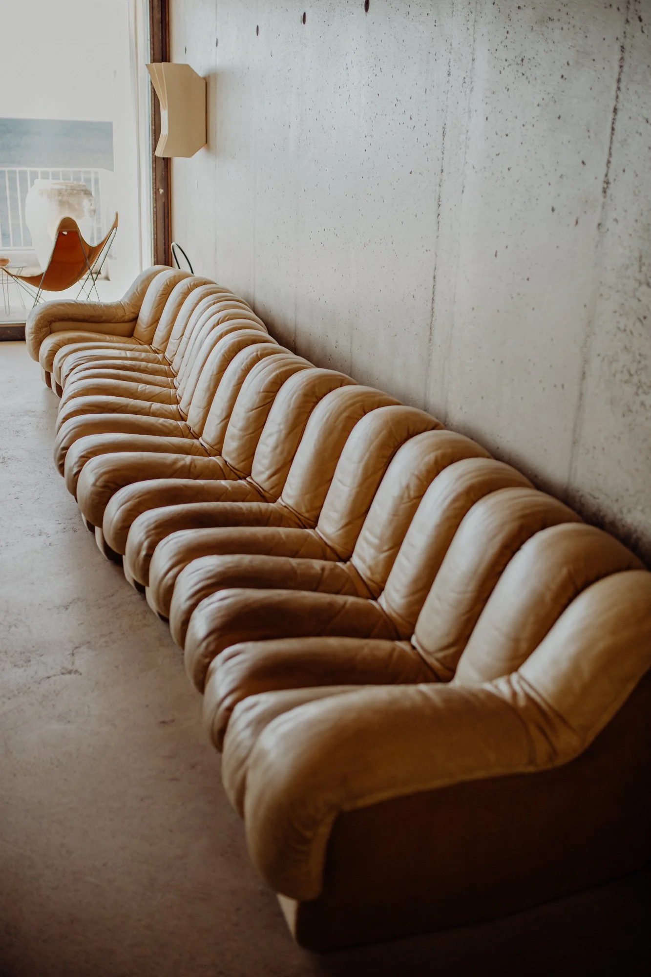 Long beige vintage sofa with tufted cushions in a room with concrete wall, beige carpet, and a window with a chair outside.