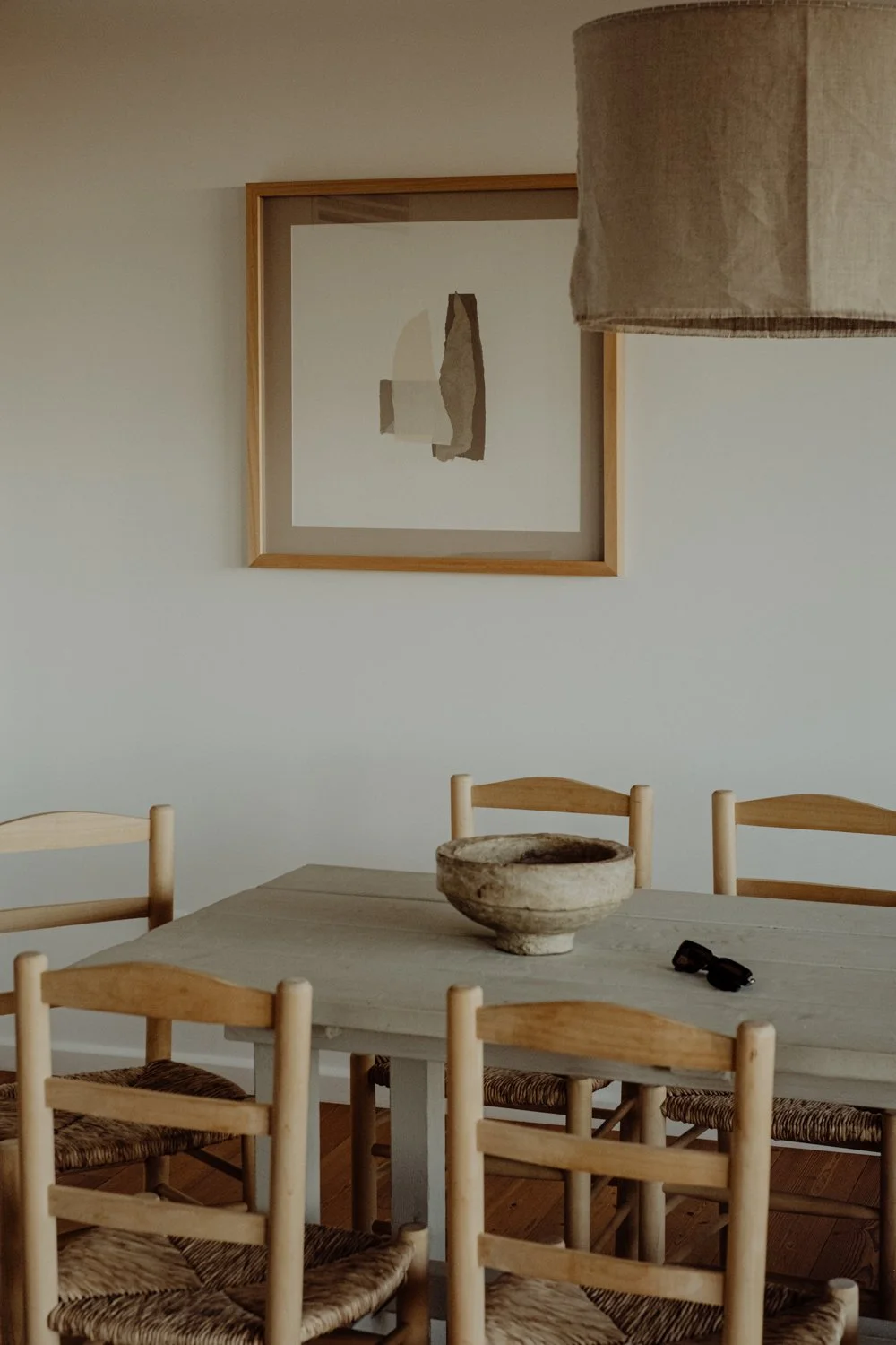A dining room with a wooden table surrounded by six chairs with woven seats, a stone bowl in the center, a pair of glasses, and a minimalist artwork on the wall.