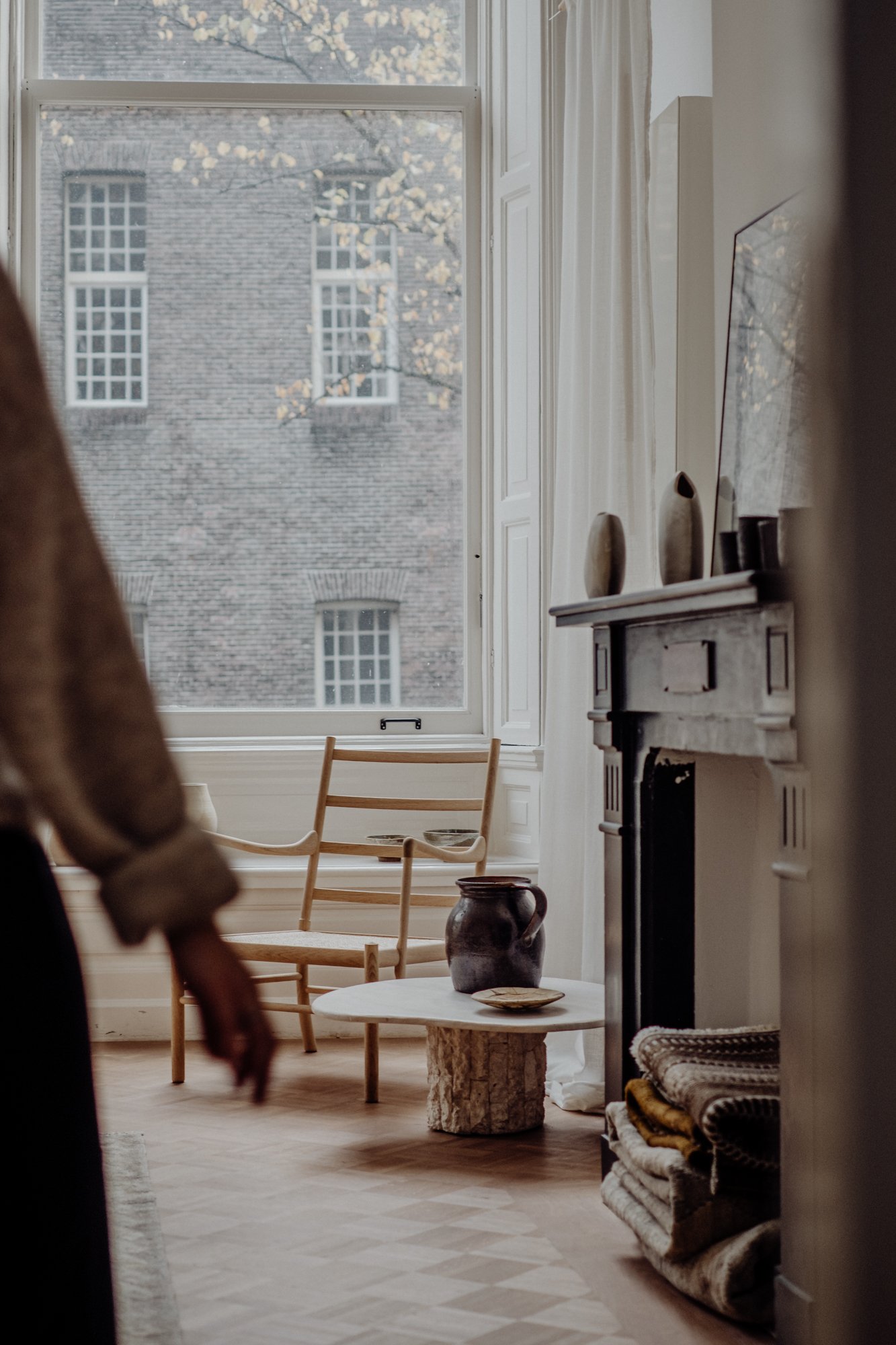Interior of a cozy room with a large window, wooden chairs, a small round table with a ceramic pitcher, and a fireplace with decorative vases on the mantel. Folded blankets are stacked nearby.