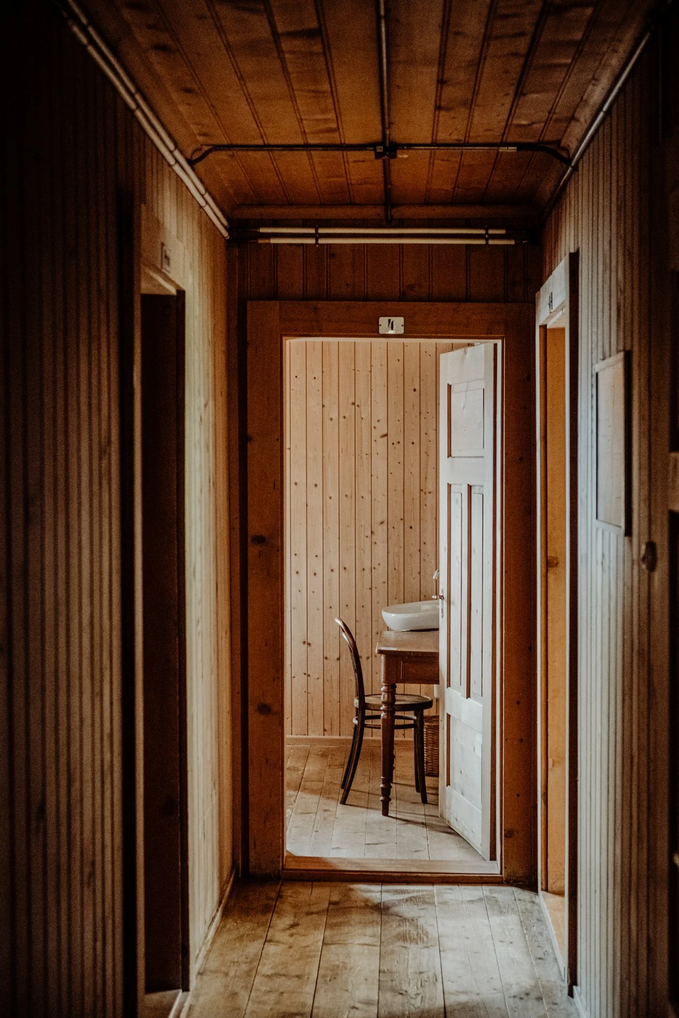 A view down a wooden hallway toward a small room with a wooden table, a black chair, and a white basin sink. The walls, ceiling, and floors are all made of wood paneling.