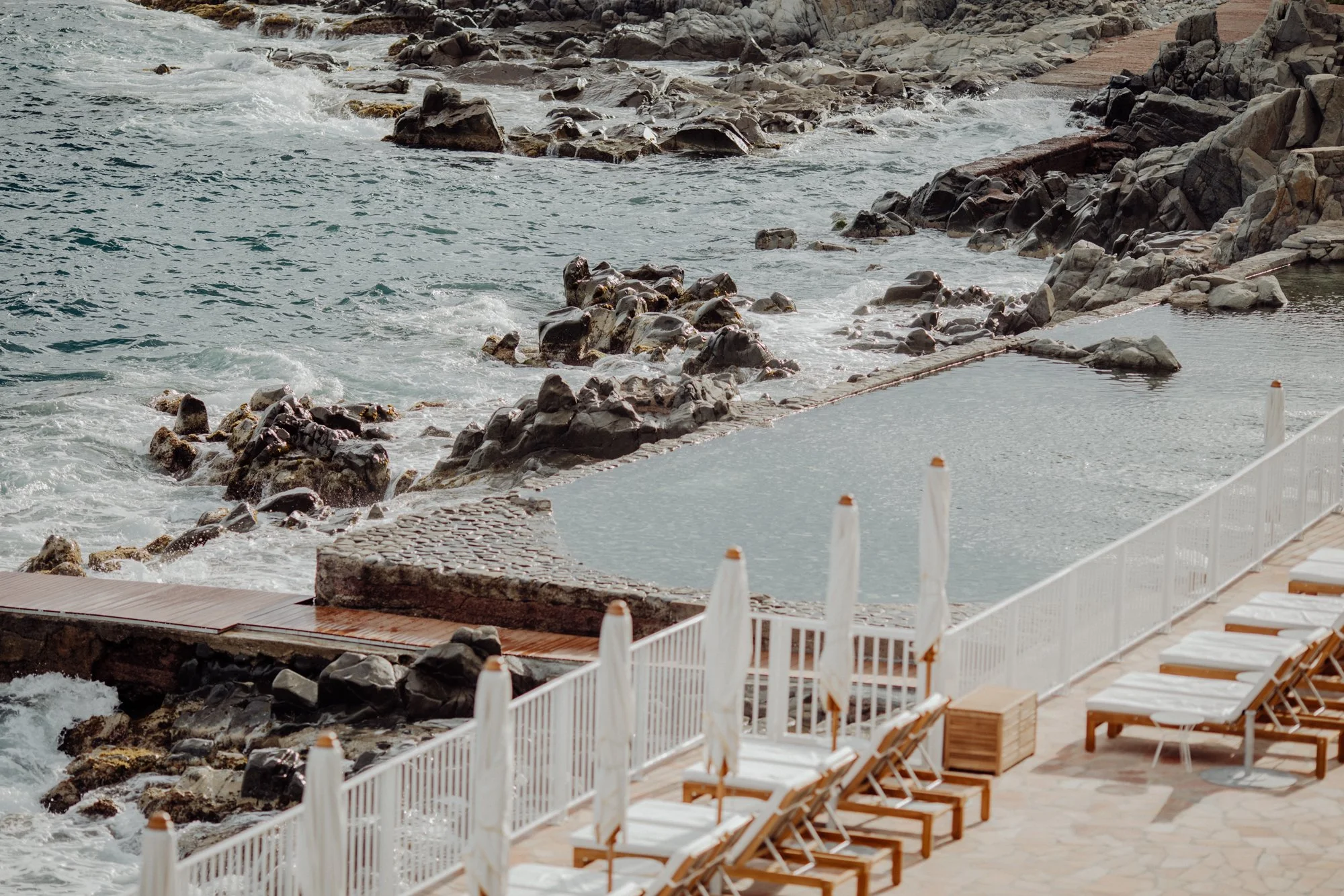 Empty poolside area with white lounge chairs and umbrellas, next to a rocky shoreline and ocean waves.