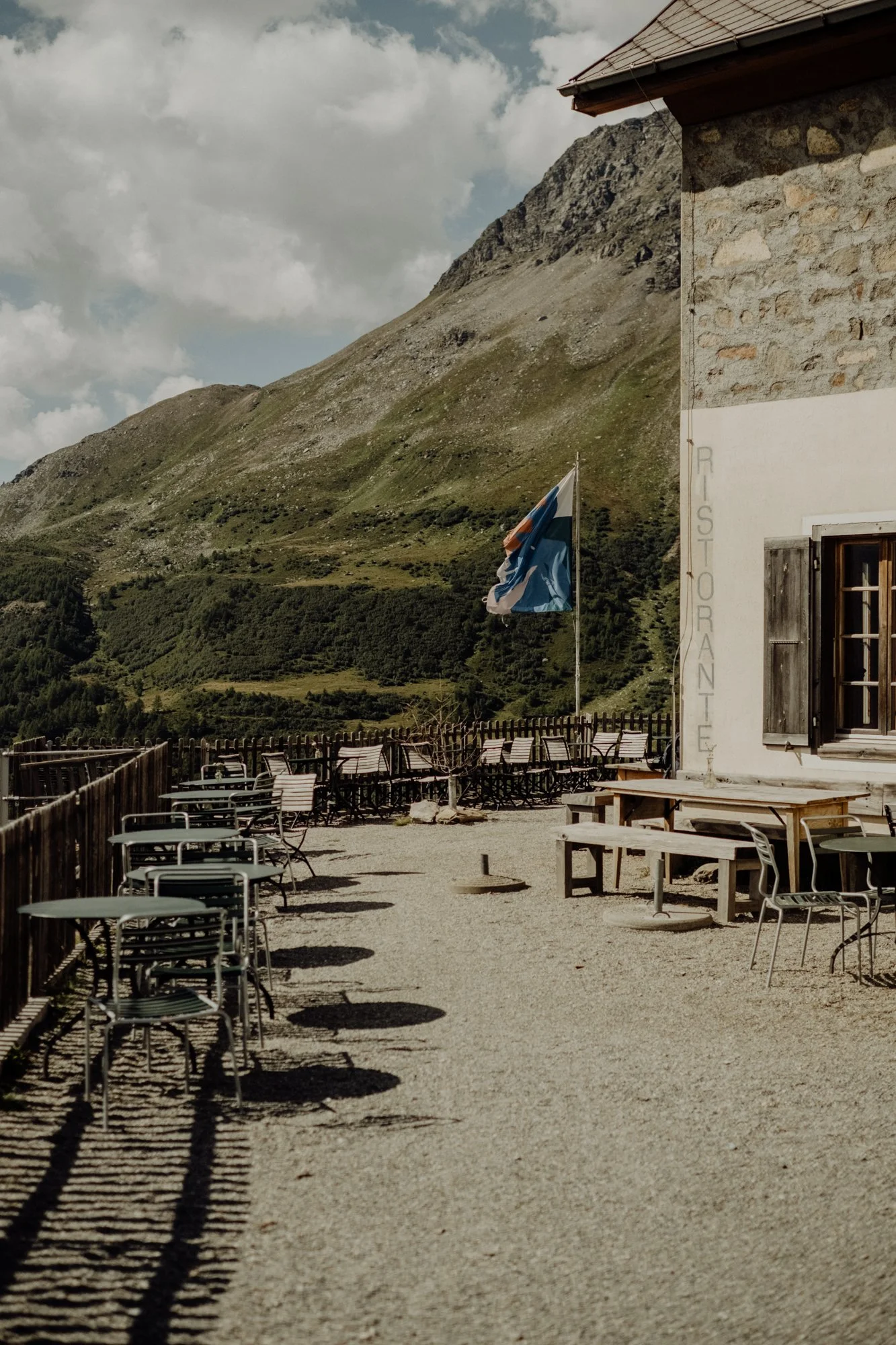 Outdoor terrace with metal chairs and tables, building with a window, a flagpole with a blue and red flag, and mountains in the background under a cloudy sky.