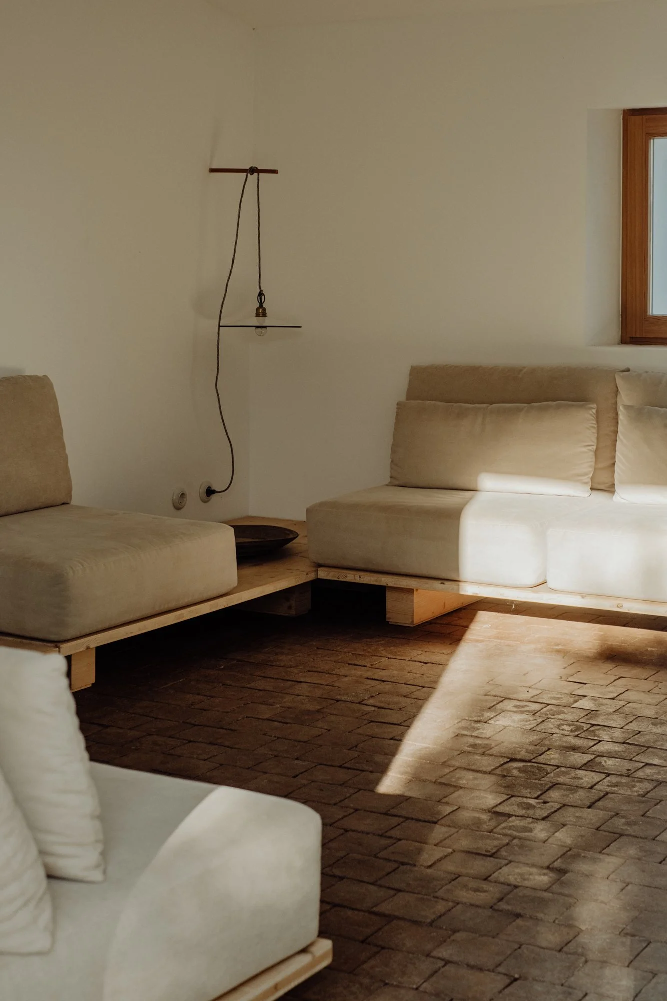 Minimalist living room with neutral-toned furniture and exposed brick flooring, featuring a beige sofa and a corner wall with a window, illuminated by natural sunlight.