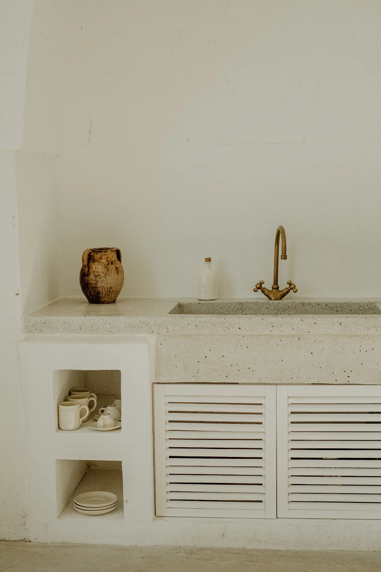 Minimalist off-white kitchen countertop with a brown vase, a small bottle, and a brass faucet, with white shelves containing cups, saucers, and plates below.