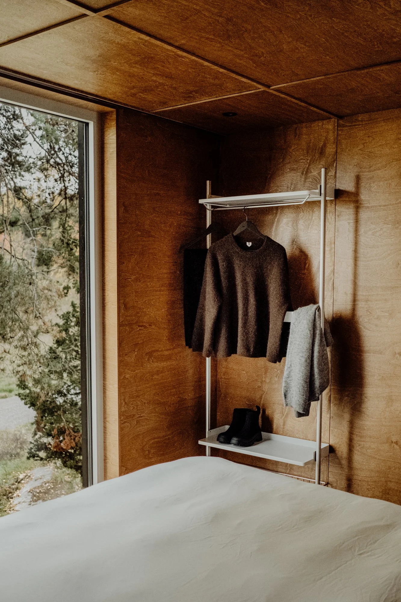 A bedroom with wooden walls and ceiling, bed with white bedding, and a standing clothing rack holding a dark sweater, a gray jacket, black shoes, and a small black item.