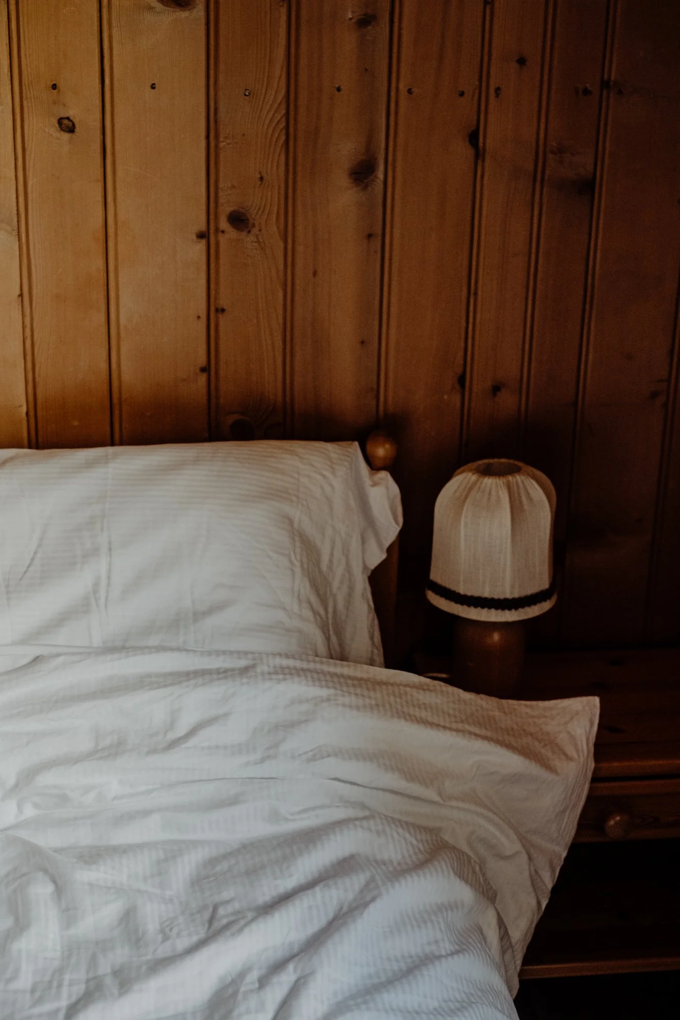 A cozy bedroom with a wooden headboard, a white pillow, and a matching white bedspread. There is a lamp with a beige shade featuring a black band, positioned on a nightstand beside the bed.