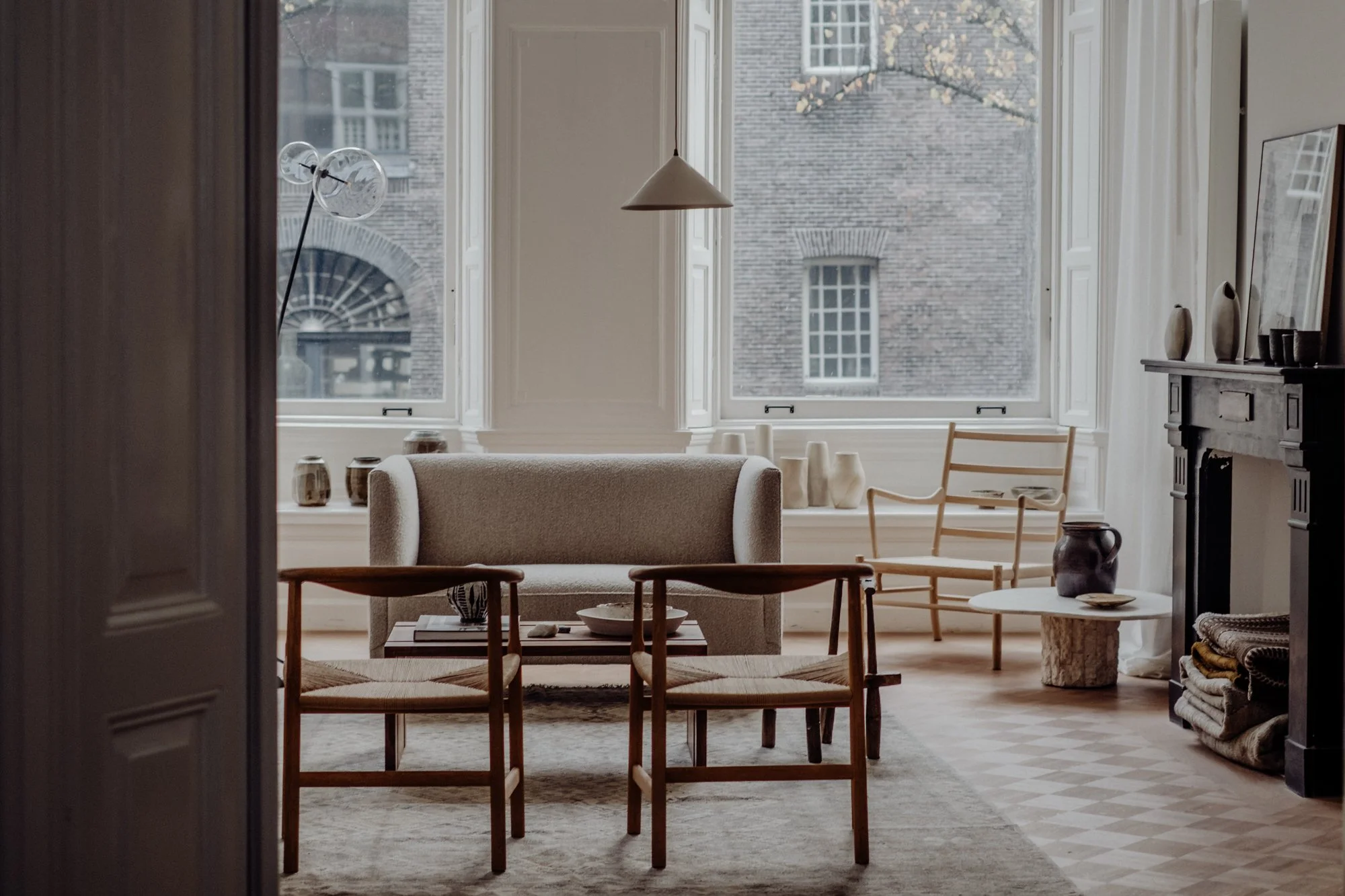 A cozy living room with beige sofa, wooden chairs, and a decorative fireplace, illuminated by natural light from large windows.