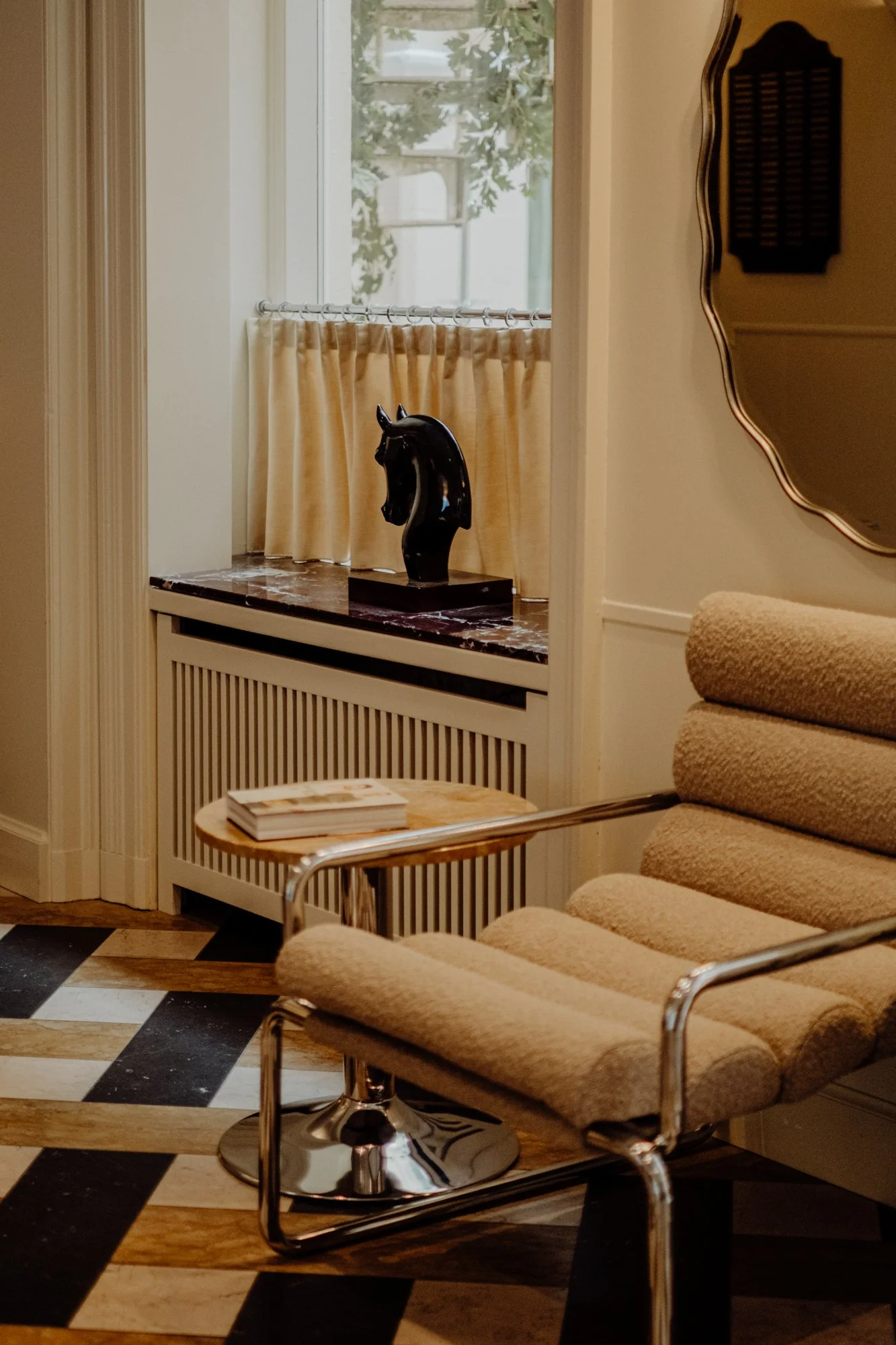Interior of a room with a beige lounge chair, a small round wooden side table with magazines, a gold-framed mirror, and a window with a horse head sculpture on the ledge.