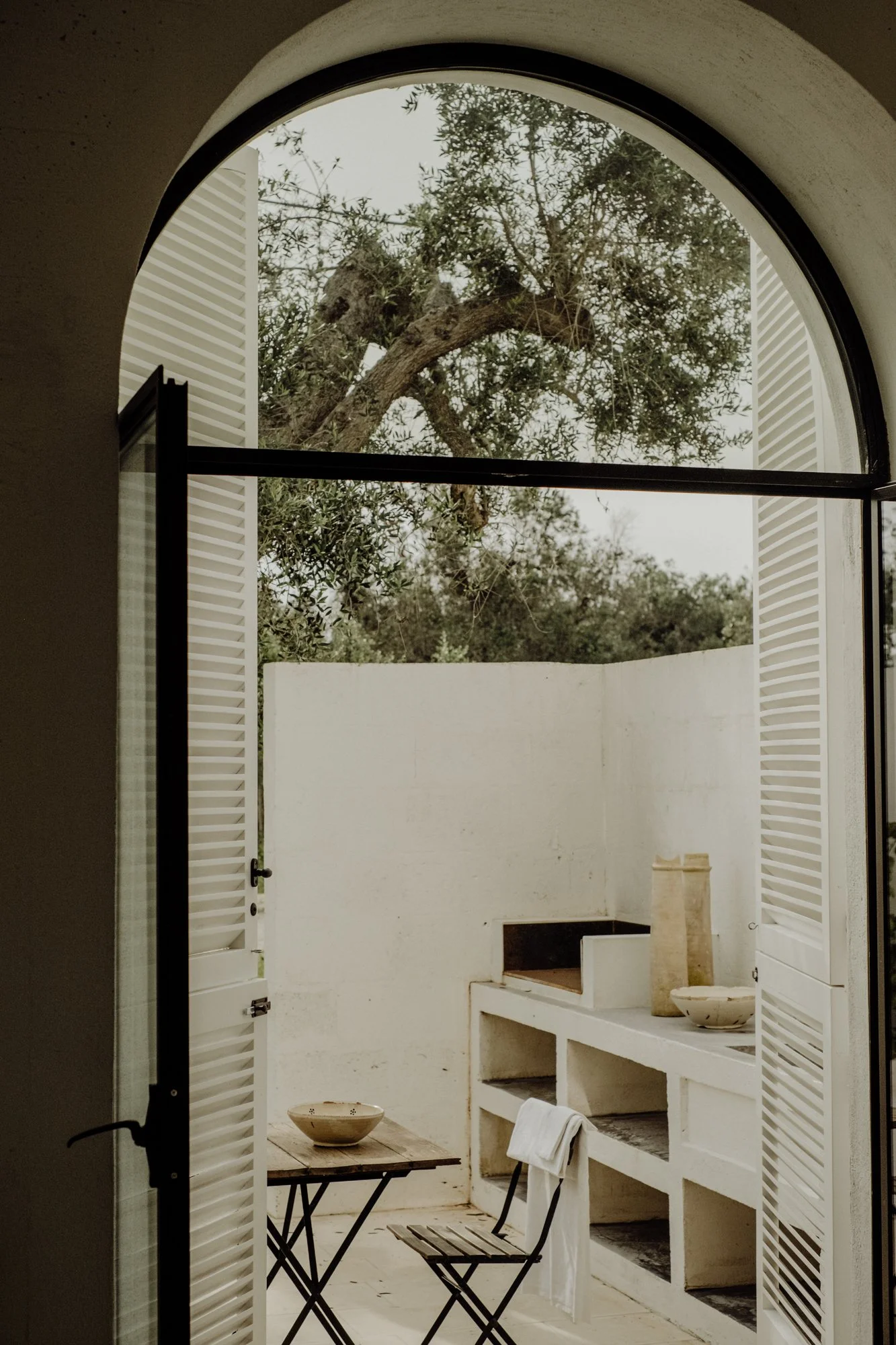 View through arched window showing a small outdoor patio with a table, chairs, and a white wall, overlooking a tree outside.
