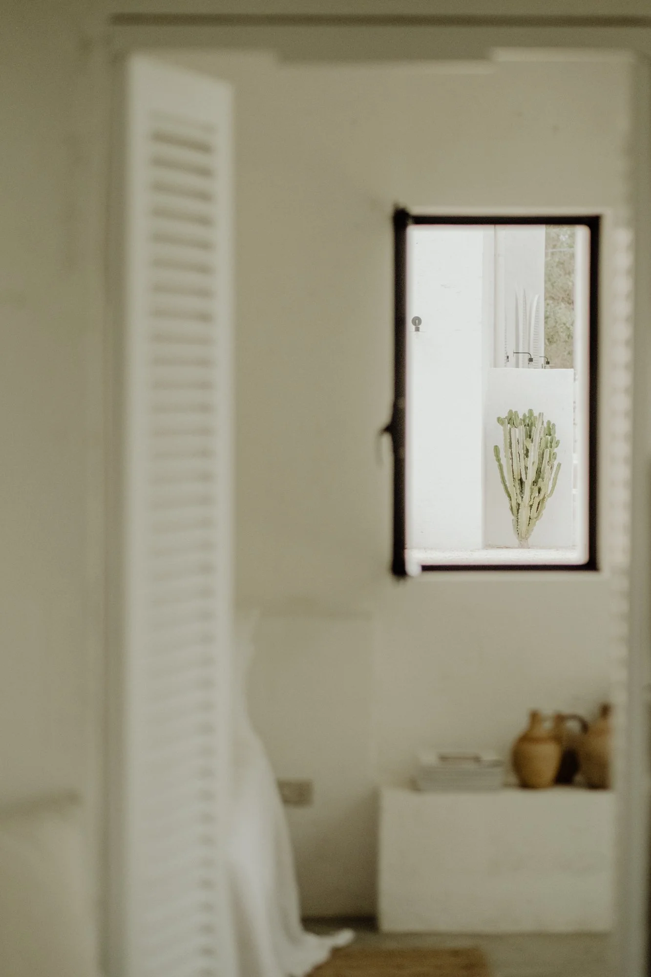 A view through a mirror showing a room with a white wall, a window with a cactus plant outside, and decor such as pottery and books inside.