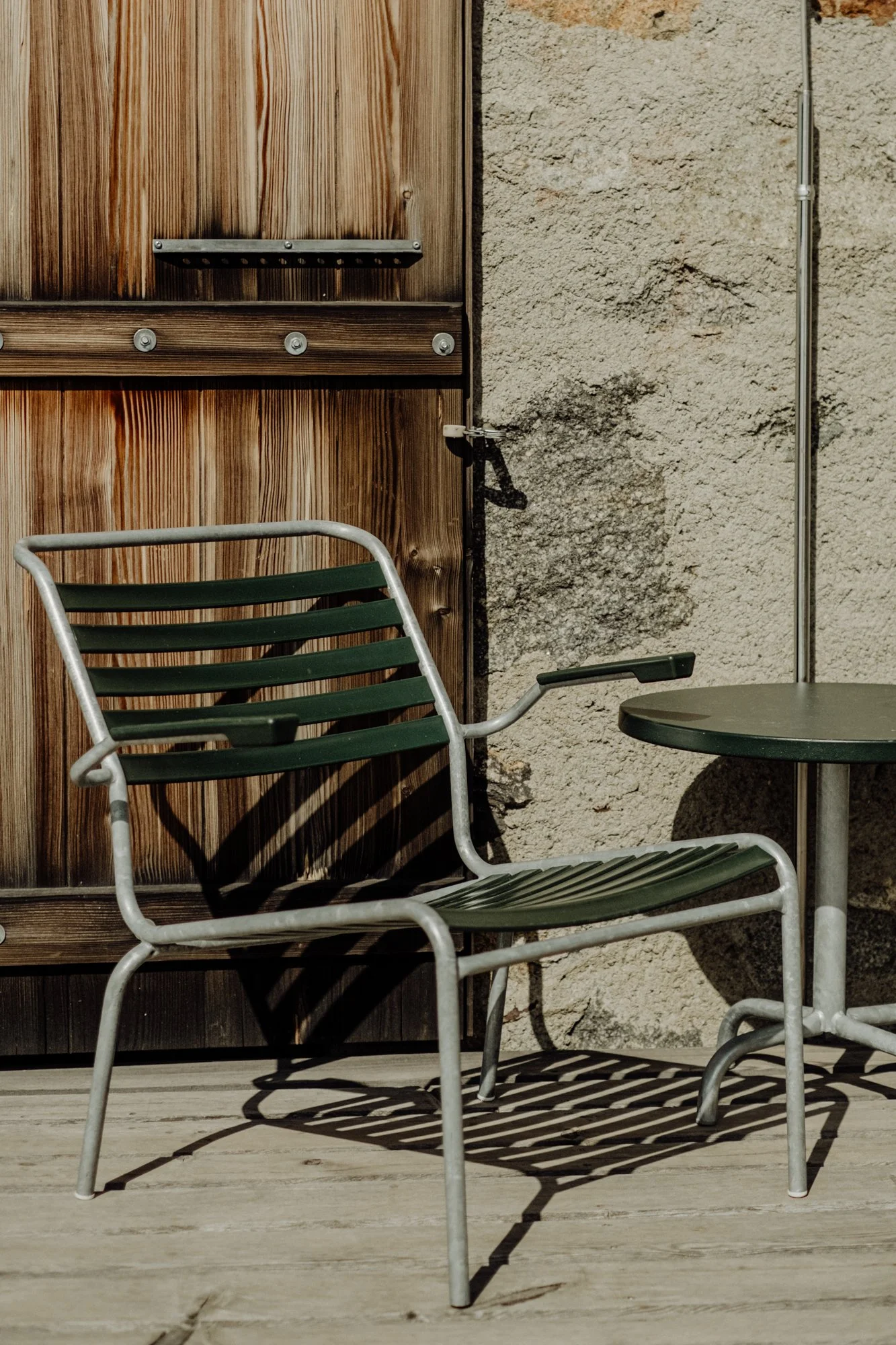 An outdoor scene with a metal and green slatted chair, a small round green table, a wooden door, and a stone wall casting a shadow.