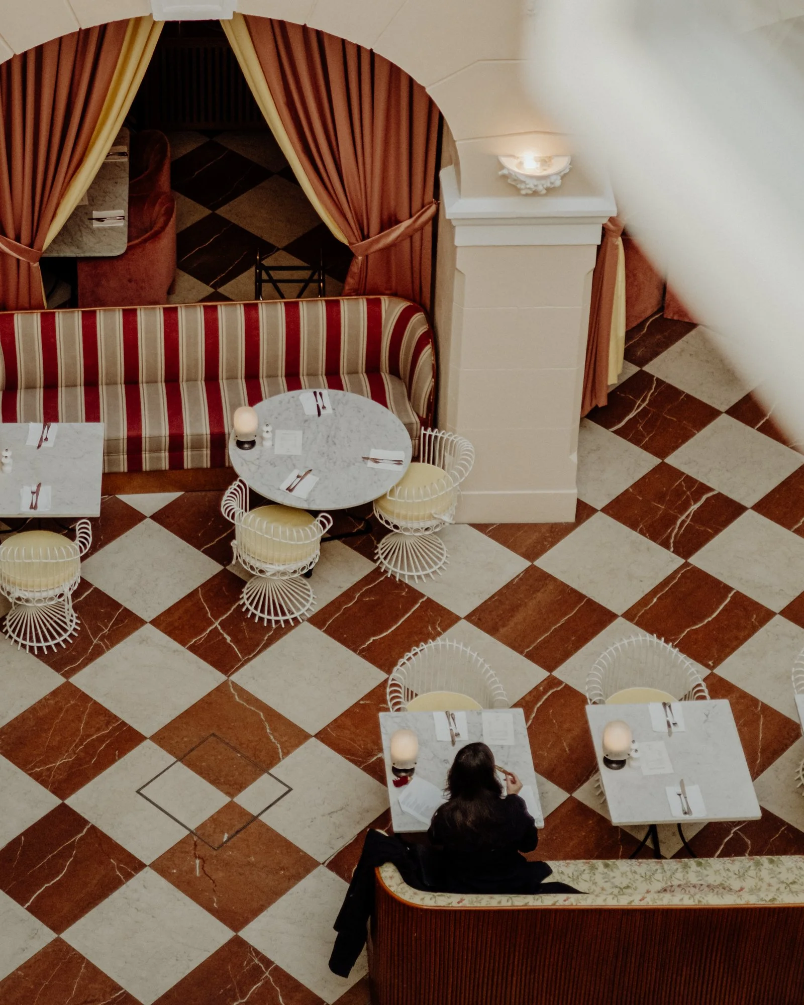 An overhead view of a restaurant dining area with checkered marble flooring, a woman sitting alone at a table, two empty tables with chairs, and a curved sofa with a marble top and pink curtains.
