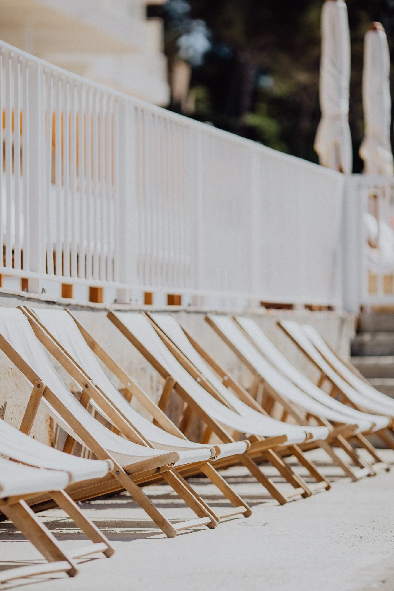 Row of empty lounge chairs beside a white pool fence, with umbrellas in the background.